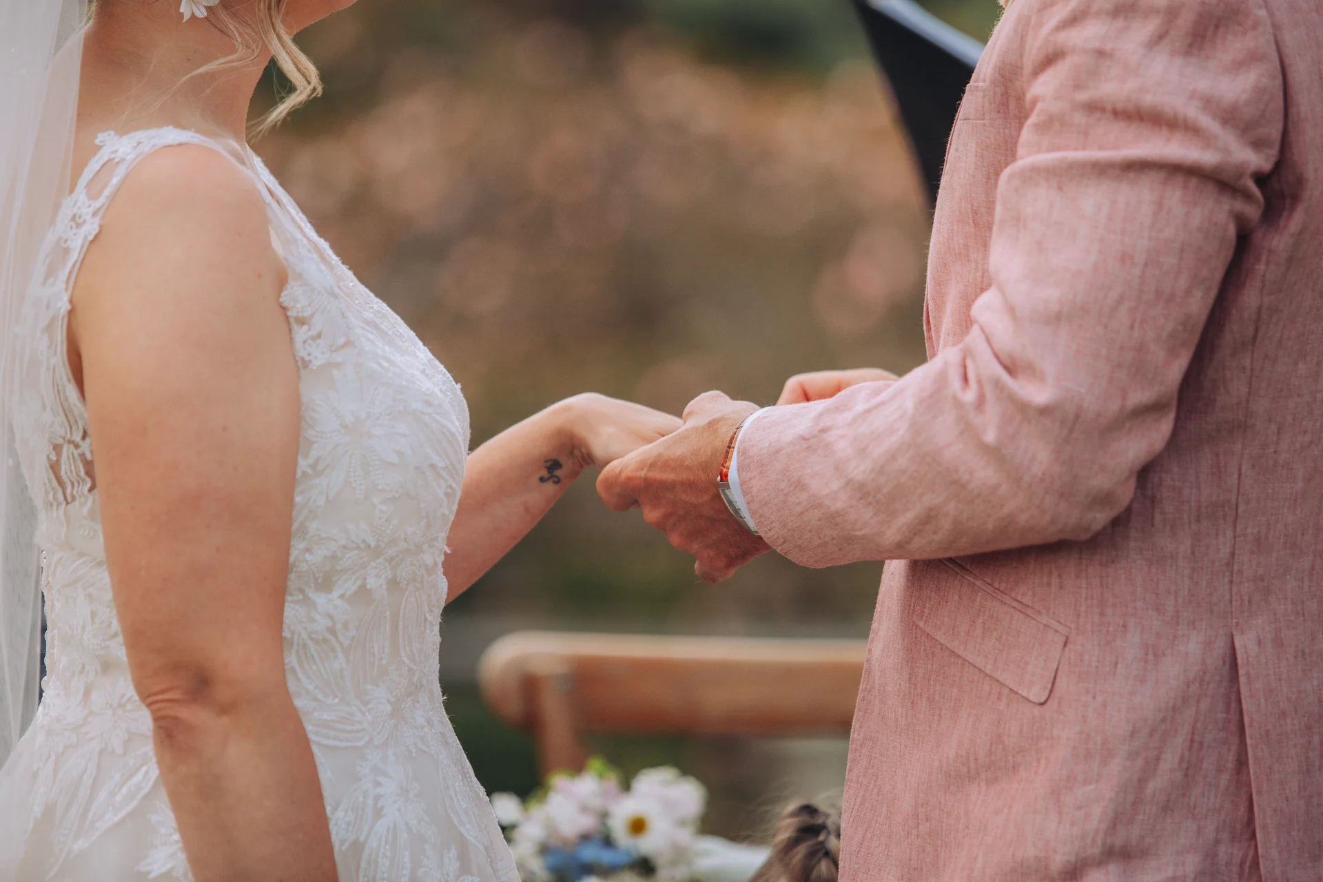 A bride in a lace wedding dress and a groom in a pink suit exchange rings during an outdoor ceremony.