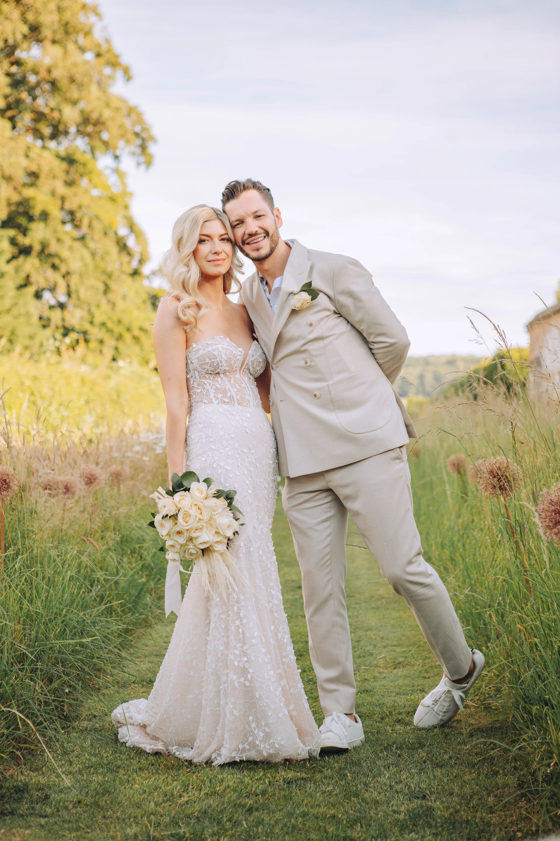 A smiling newlywed couple poses together in a lush green field, with the bride holding a white rose bouquet and wearing a strapless lace gown while the groom wears a cream suit and trainers.