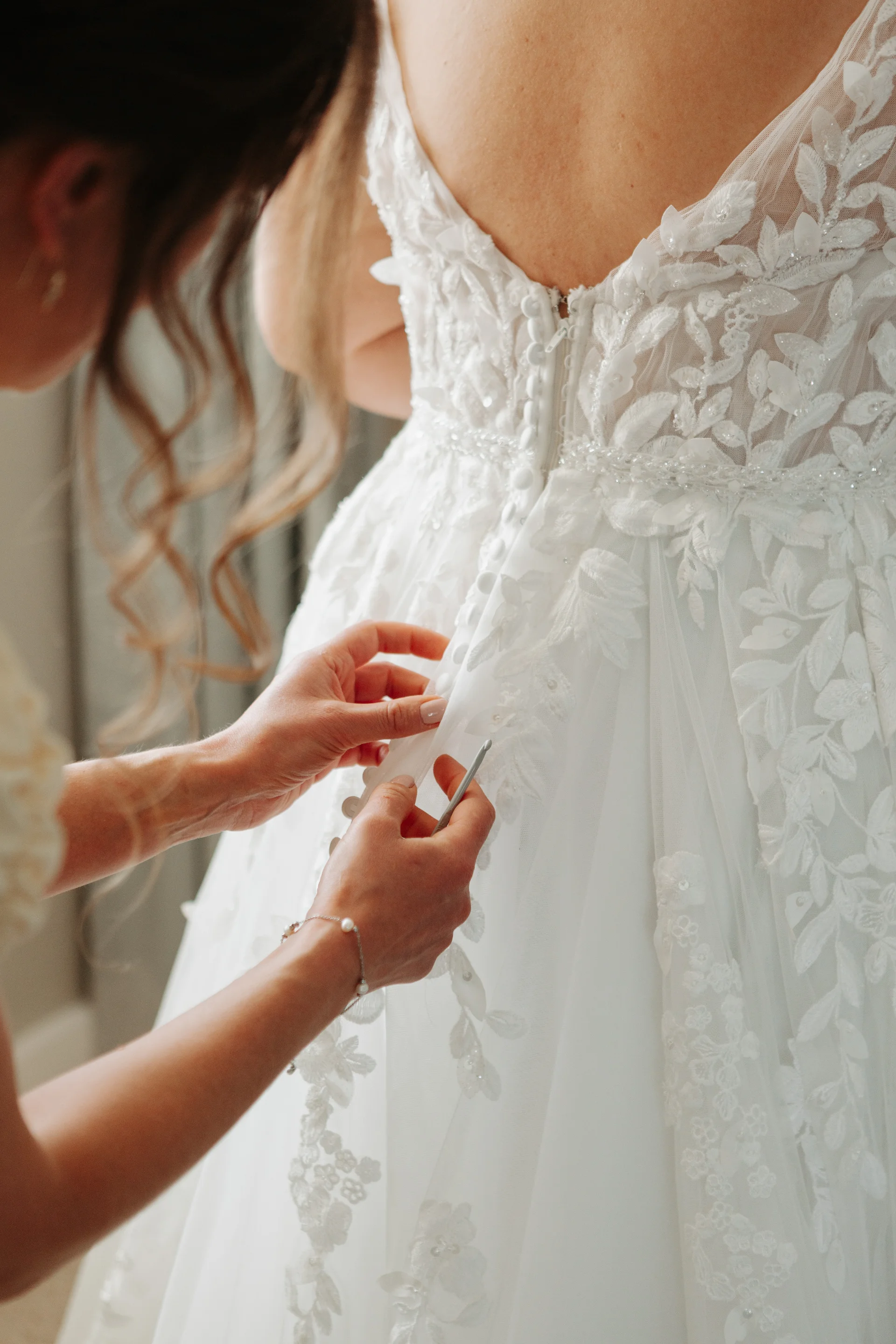 A close-up shot captures the hands of someone carefully fastening the intricate button back of a detailed lace wedding dress.