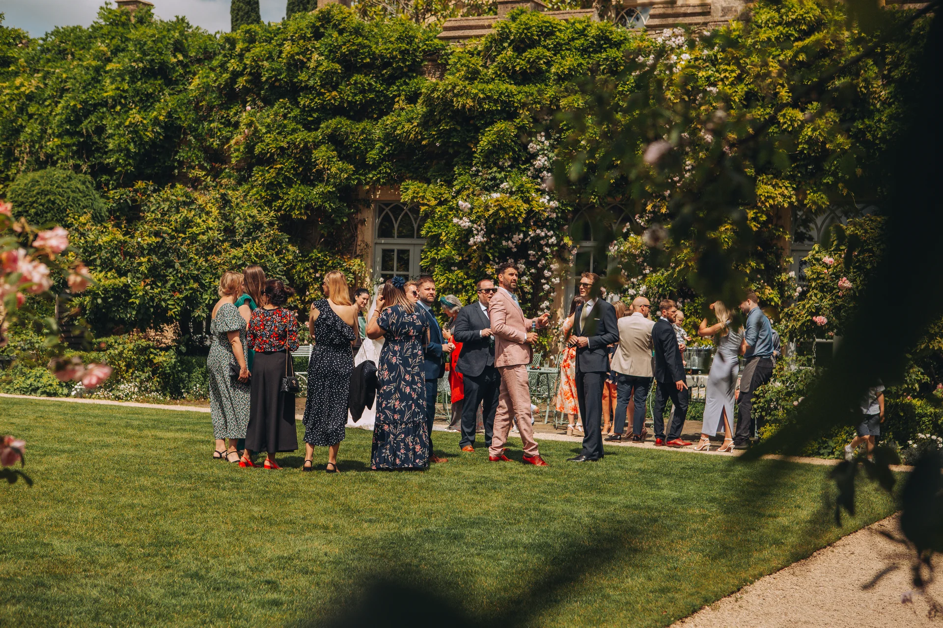 Wedding guests mingle and chat on a lush green lawn during a sunny drinks reception in the walled garden of a country house.