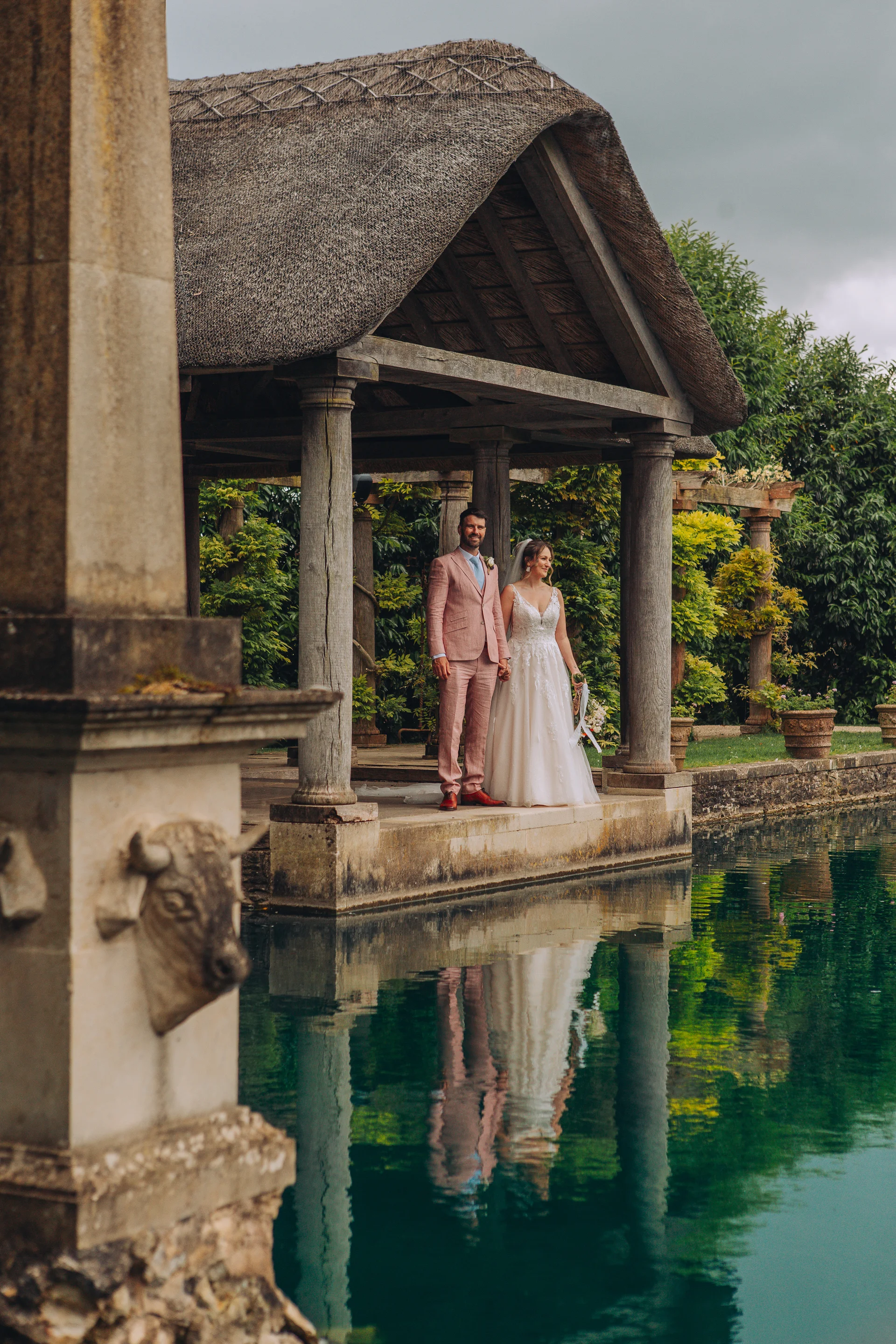 A bride in an a-line gown and a groom in a pink suit stand hand in hand under a thatched lakeside gazebo, perfectly reflected in the water below.