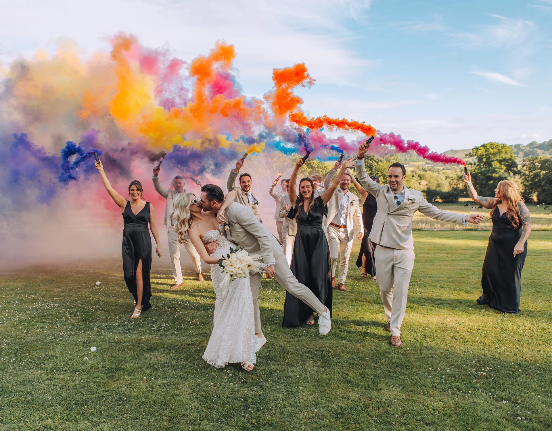 A newlywed couple share a dramatic dip kiss in the foreground on a lush green lawn, while their wedding party enthusiastically sets off vibrant clouds of colourful smoke bombs behind them under a bright blue sky.
