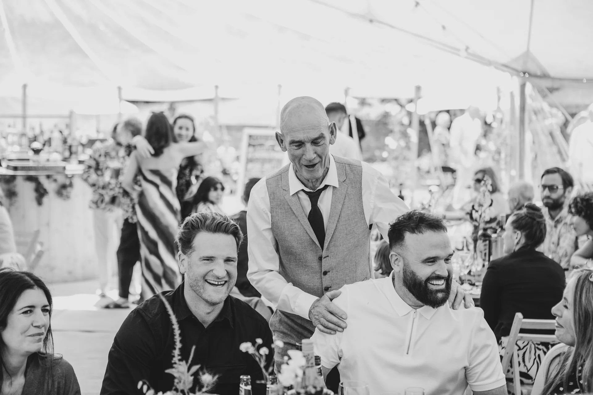 An older gentleman affectionately places his hands on the shoulders of two smiling guests during a joyous wedding reception inside a sunlit marquee.