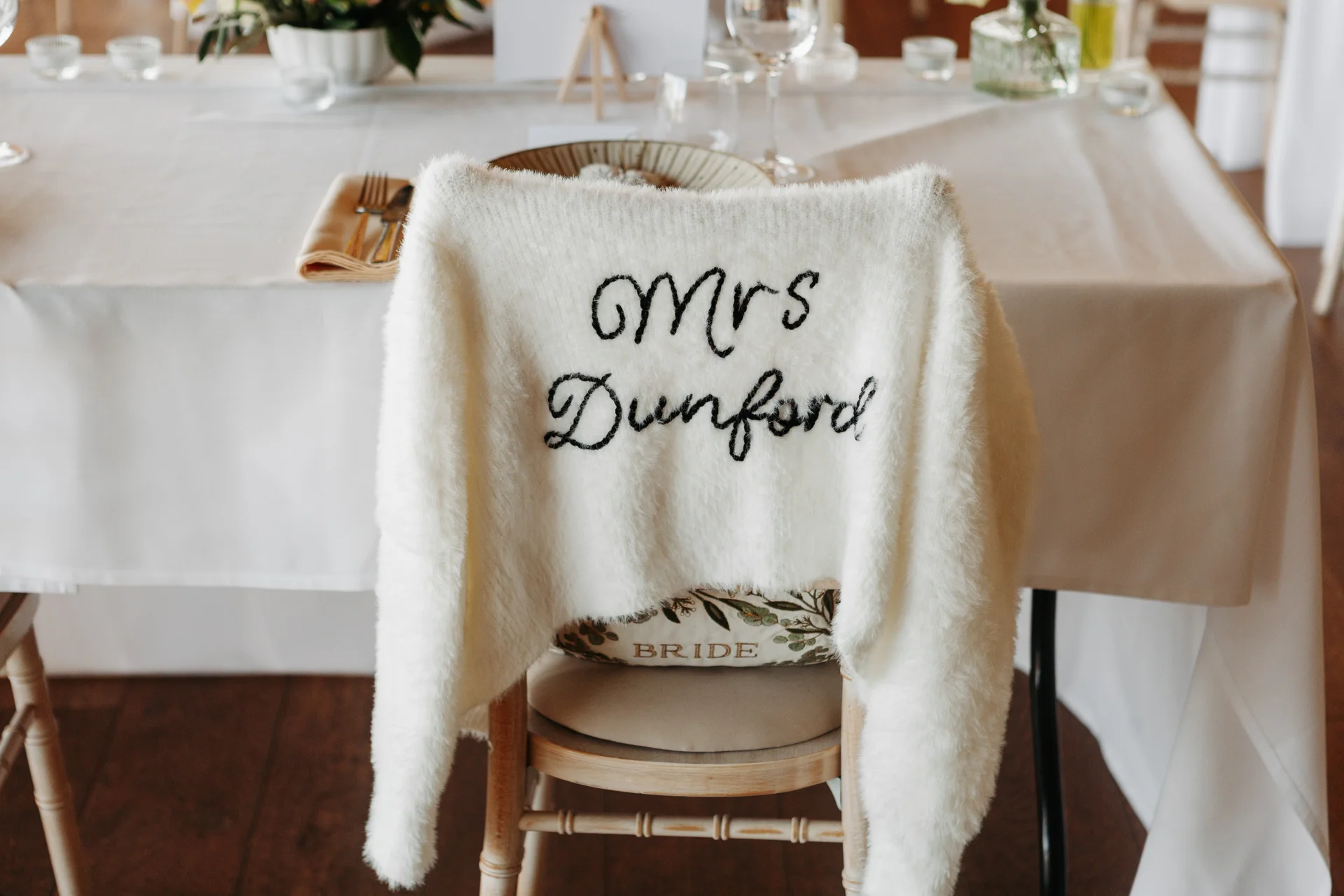 A soft white knitted cover up reading Mrs Dunford is draped over the back of a wooden wedding chair at a beautifully set reception table.