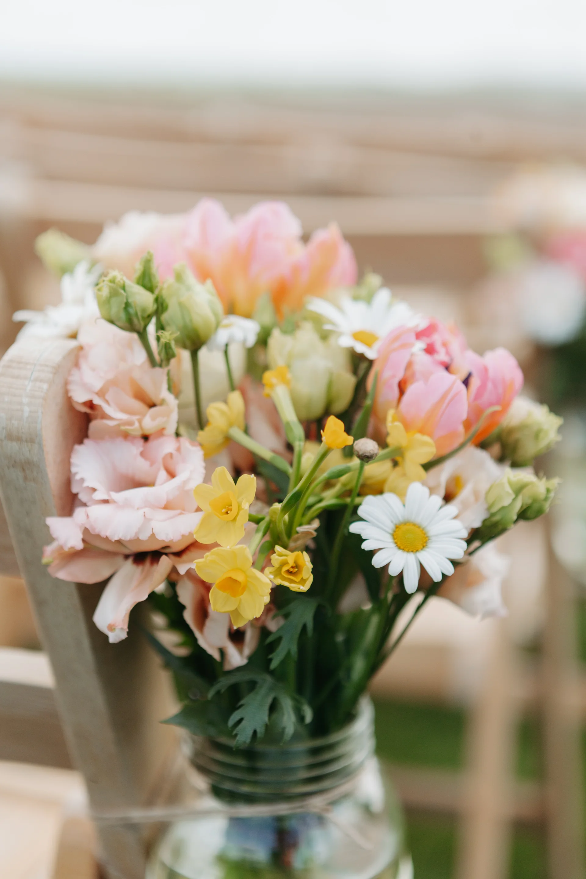 A charming spring wedding pew end features a delicate glass jar filled with colourful pastel blooms including daisies, daffodils, tulips and lisianthus.
