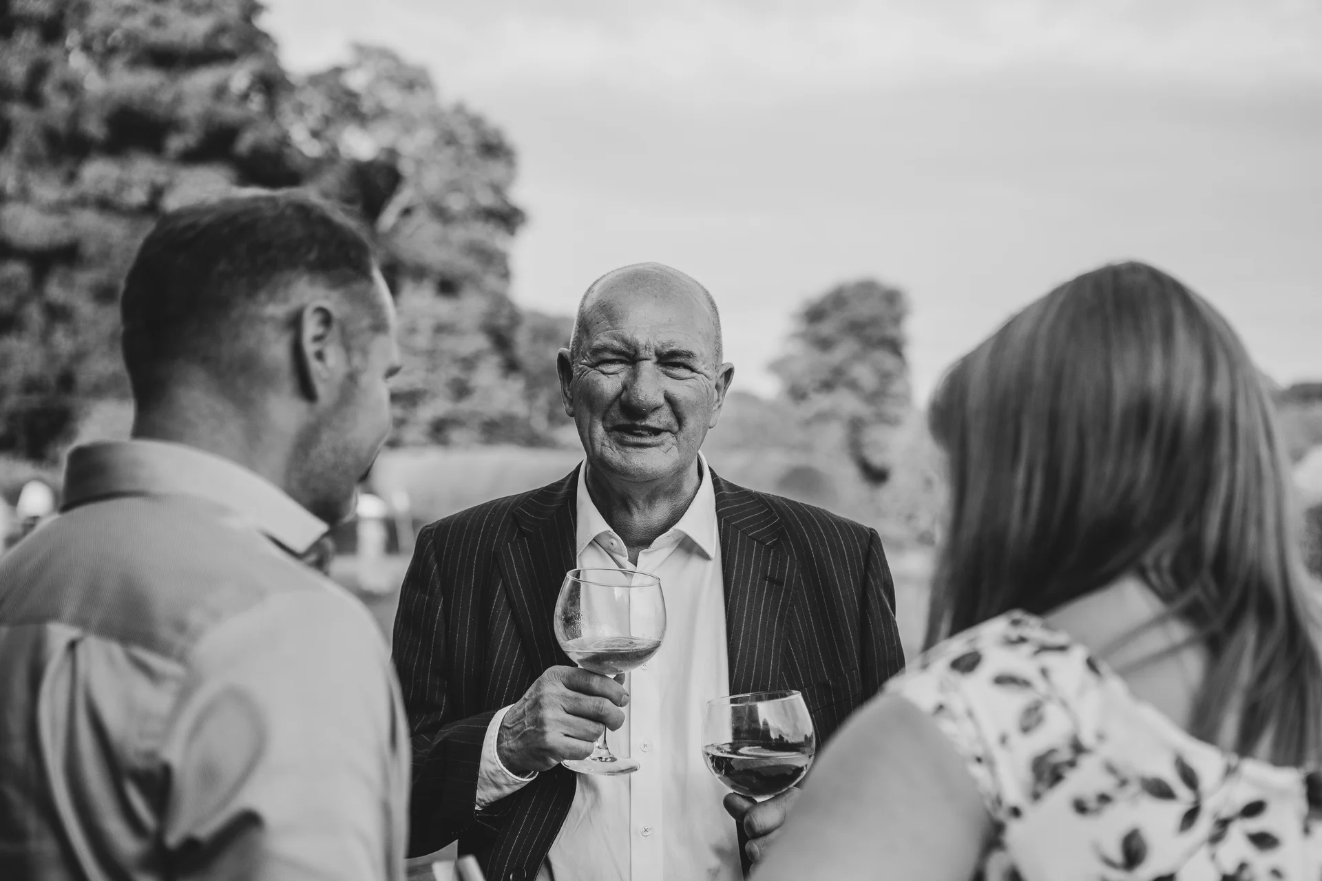 An older gentleman holding a wine glass stands outdoors and converses with wedding guests during a drinks reception.