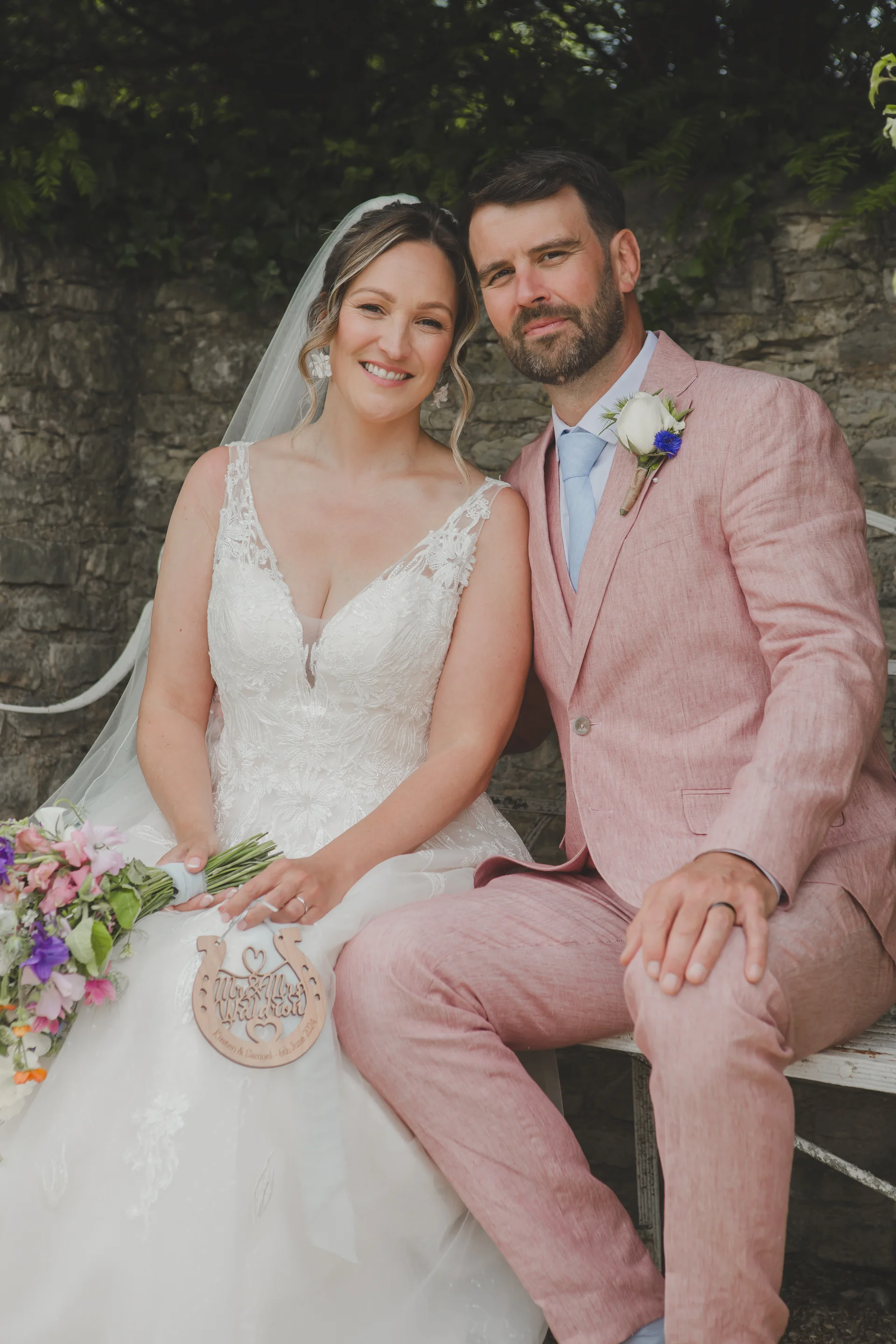 A smiling bride and groom sit together against a rustic stone wall, with the bride holding a colourful bouquet and a wooden keepsake.