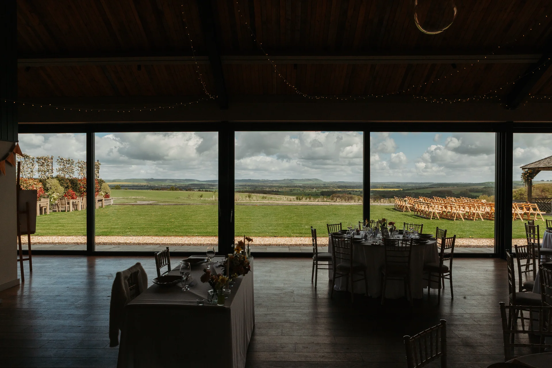 A wedding reception venue interior featuring large floor to ceiling glass windows looking out over a green lawn and rolling countryside hills, with tables set up for the wedding breakfast.