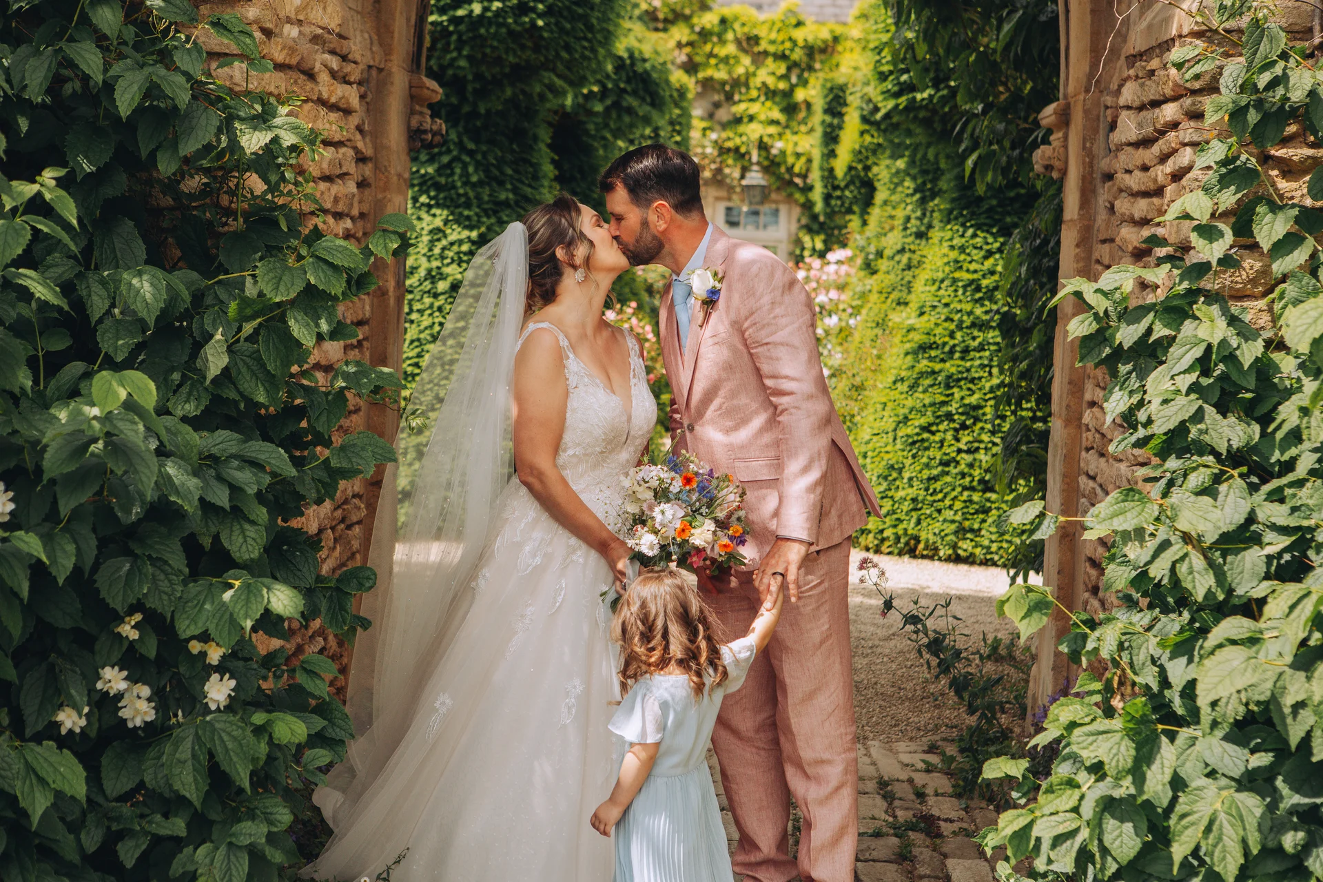 A bride in an a-line gown and veil shares a kiss with a groom in a pink suit while holding hands with a flower girl under a stone archway draped in lush greenery.