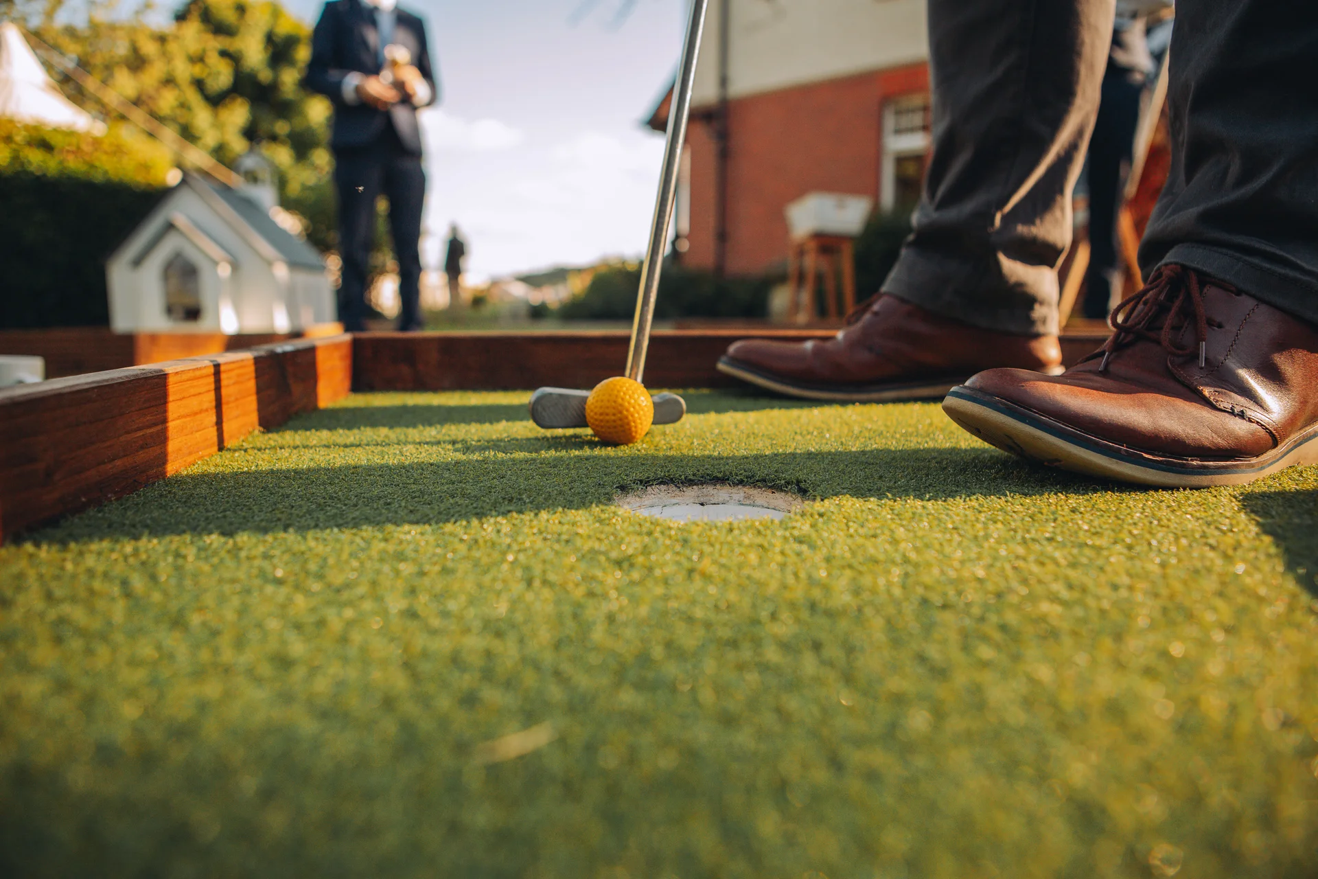 A low-angle close-up shot captures a guest playing crazy golf on an artificial lawn during a wedding reception in the golden hour sunshine.