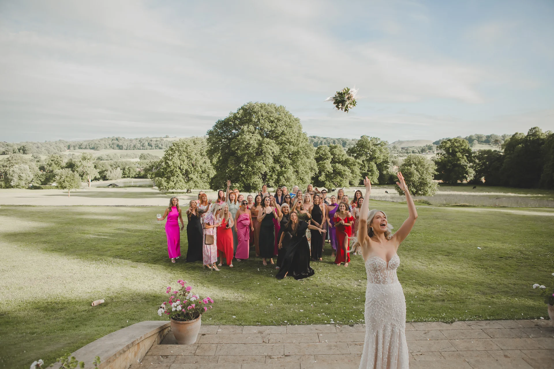 A joyful bride throws her bouquet back towards a large group of cheering guests in colourful dresses on a manicured lawn overlooking rolling hills.