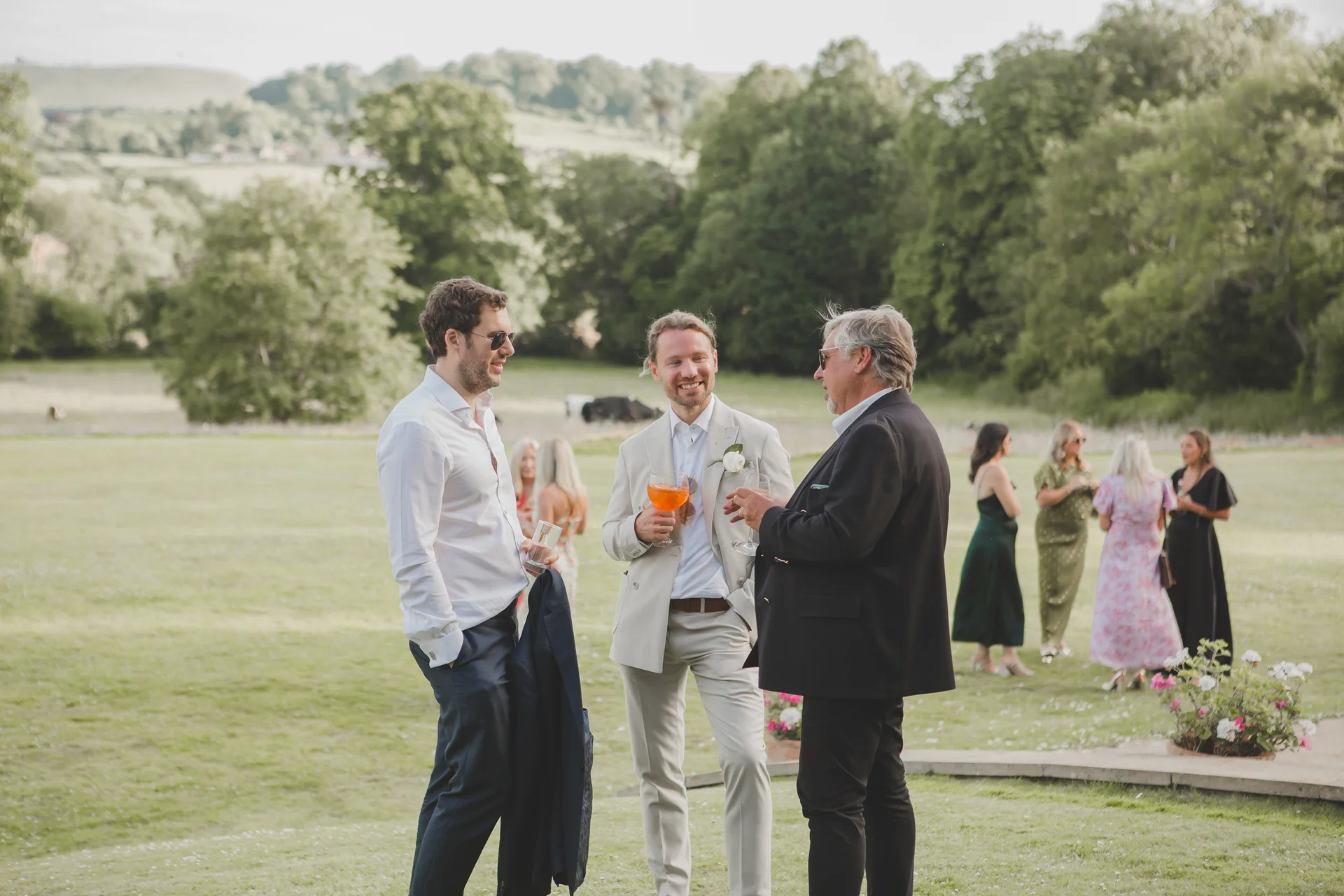 Guests enjoy drinks and conversation outdoors on a grassy lawn during a summer wedding drinks reception with rolling hills in the background.