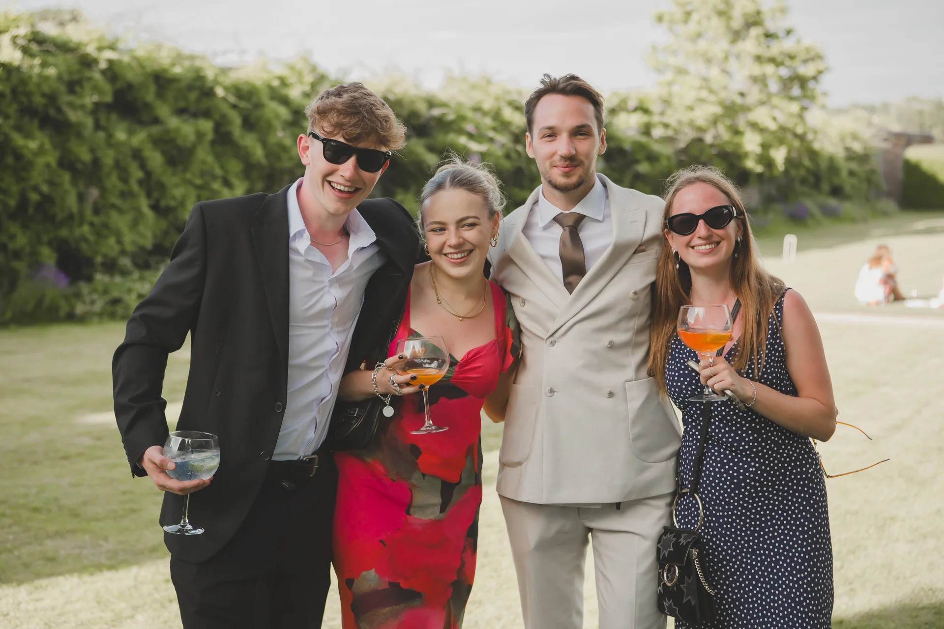 A group of smartly dressed wedding guests wearing sunglasses pose outdoors with drinks in hand during a sunny garden reception.