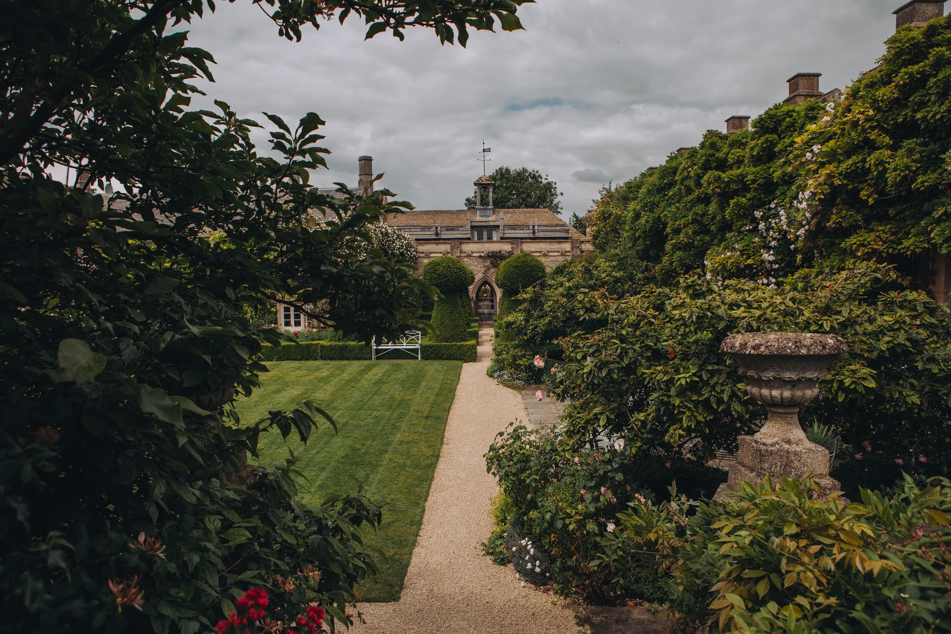 An atmospheric view of a manicured English country garden with a gravel pathway leading towards a historic manor house under a moody overcast sky.