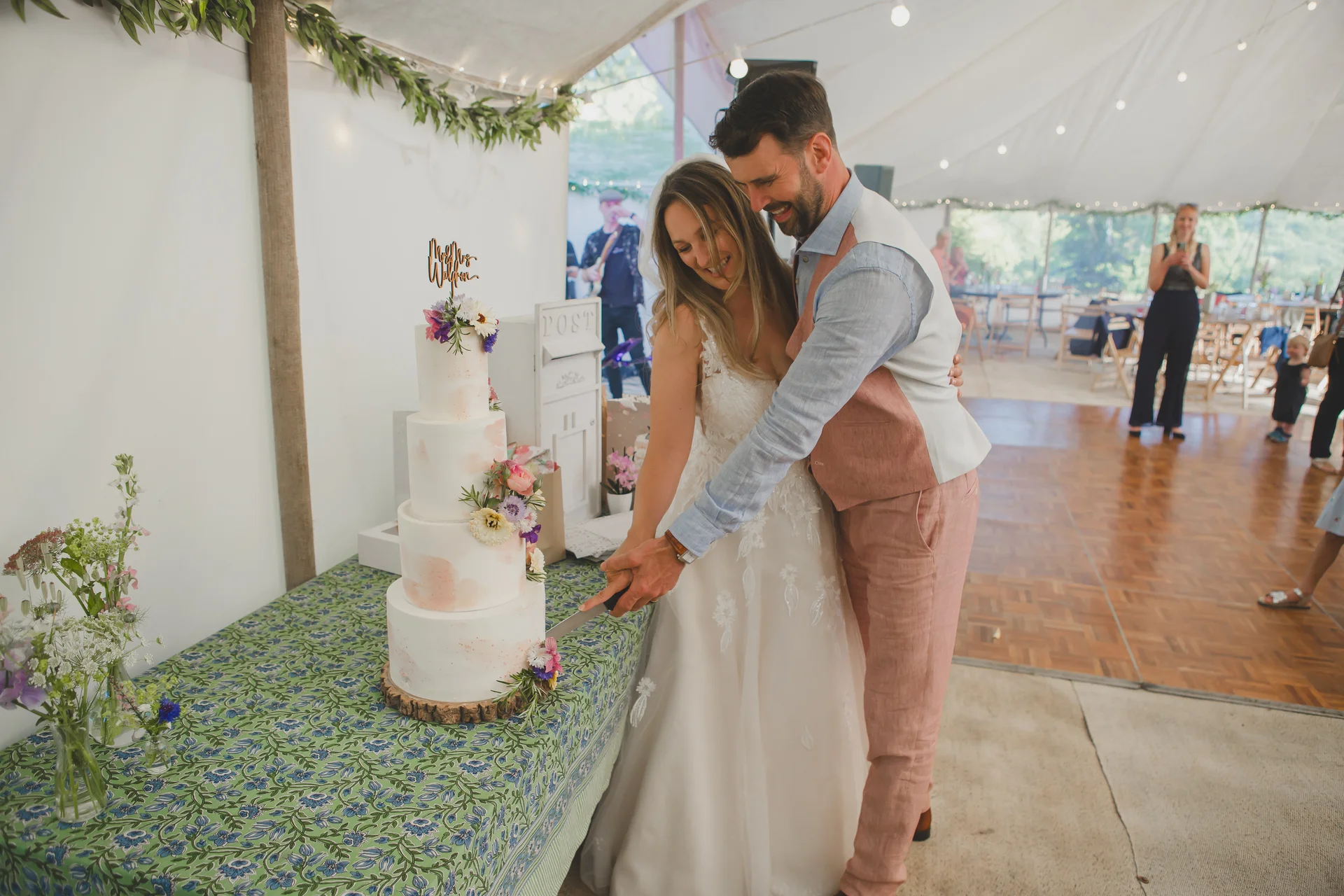 A smiling bride and groom cut a four-tier white wedding cake decorated with fresh flowers inside a brightly lit marquee.