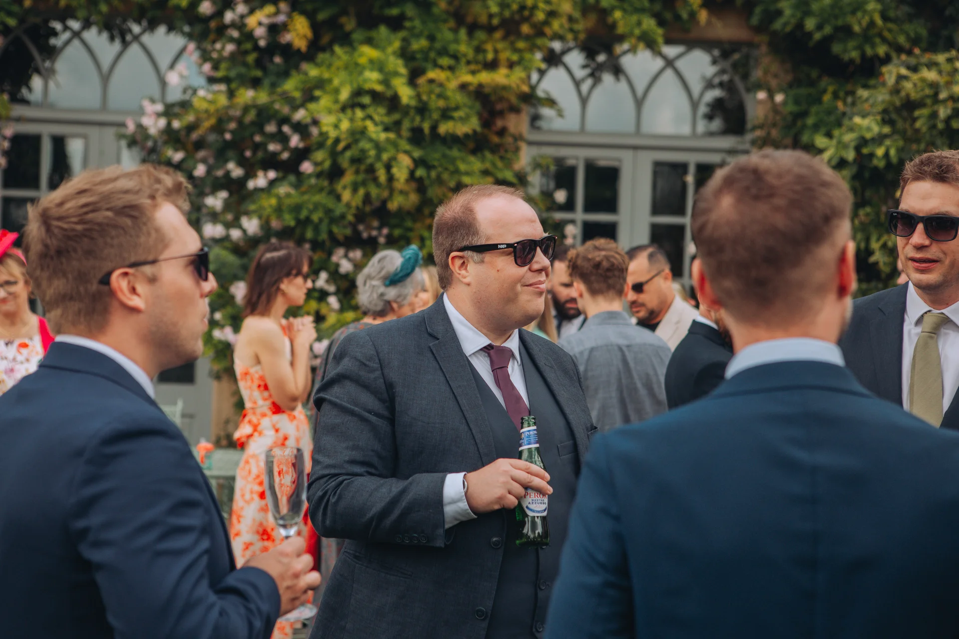 Wedding guests in suits and sunglasses mingle outdoors during a drinks reception, holding bottles and glasses near a stone building covered in climbing roses.