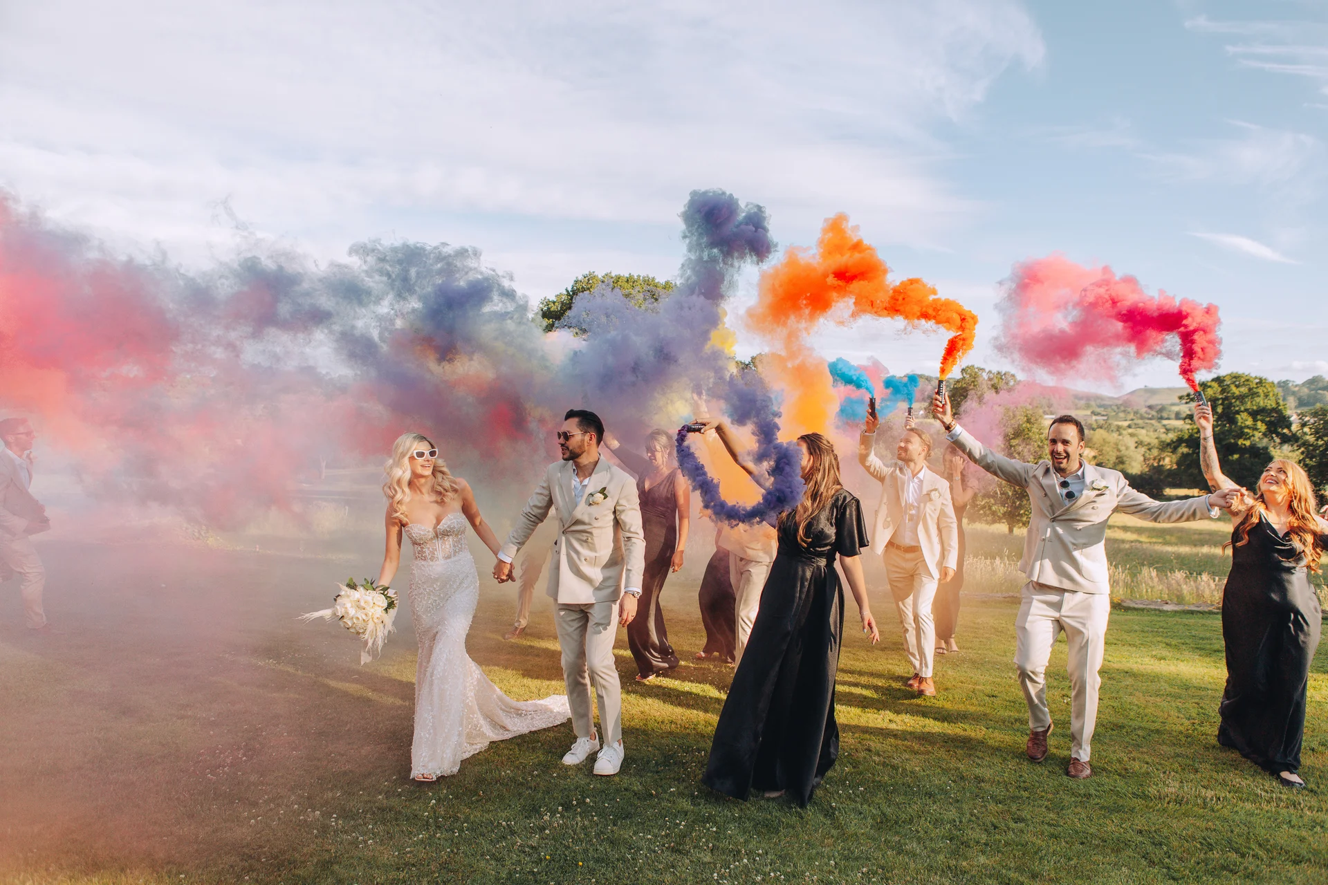 A bride and groom walk through a grassy field hand in hand while their wedding party sets off vibrant multi-coloured smoke grenades behind them.