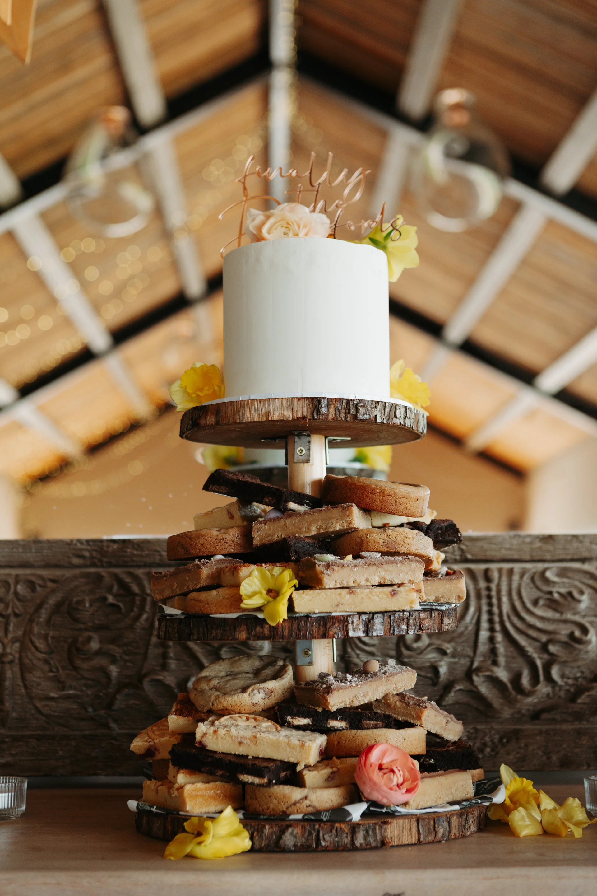A multi-tiered rustic dessert display featuring a white wedding cake on top and layers of brownies and flapjacks on wooden slice stands inside a barn.