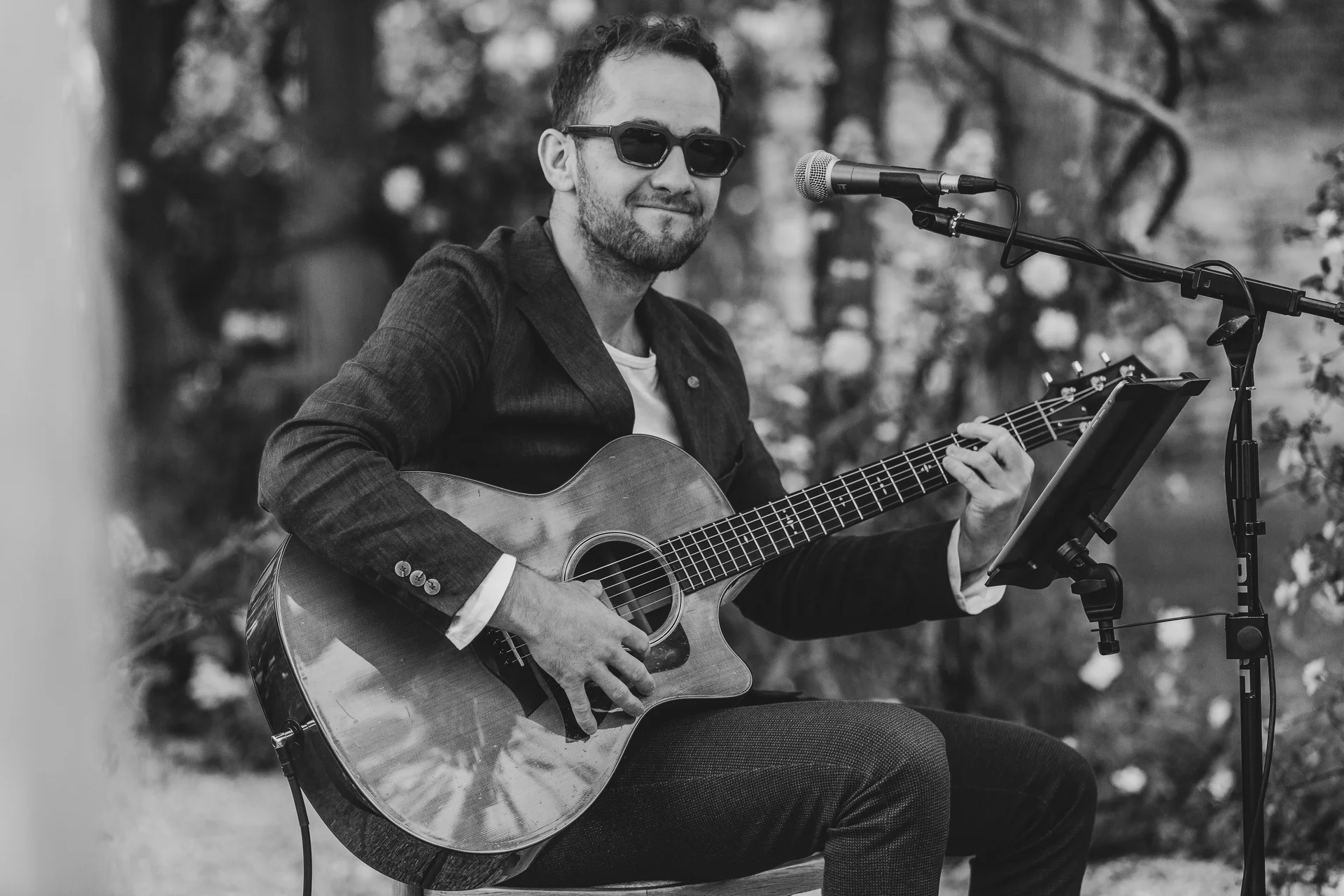A stylish male musician wearing sunglasses and a blazer performs live outdoors, playing an acoustic guitar and singing into a microphone during a wedding celebration.