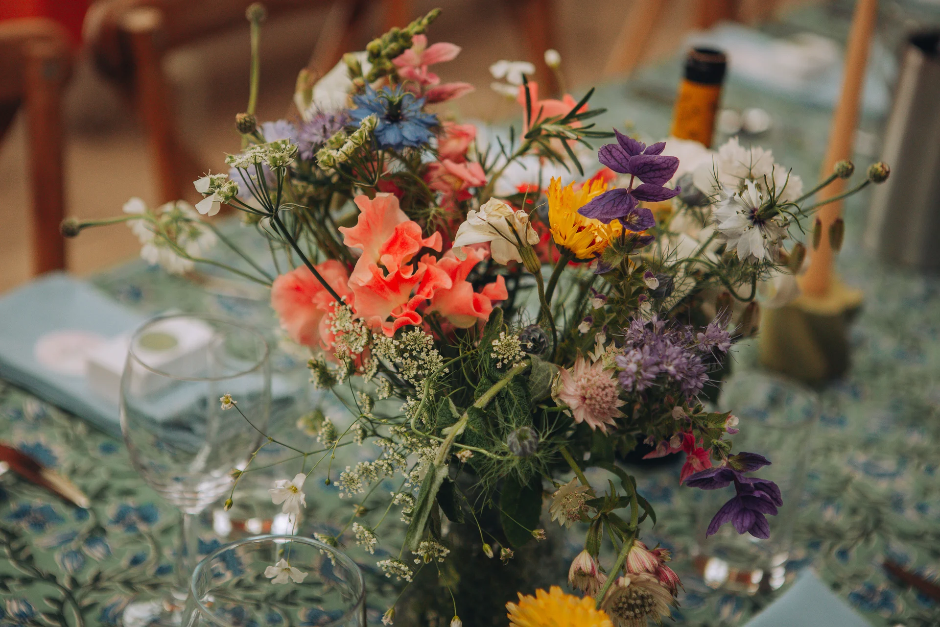 A vibrant and colourful wildflower table centrepiece featuring delicate cosmos and scabiosa arranged in a loose, organic style on a patterned linen tablecloth.