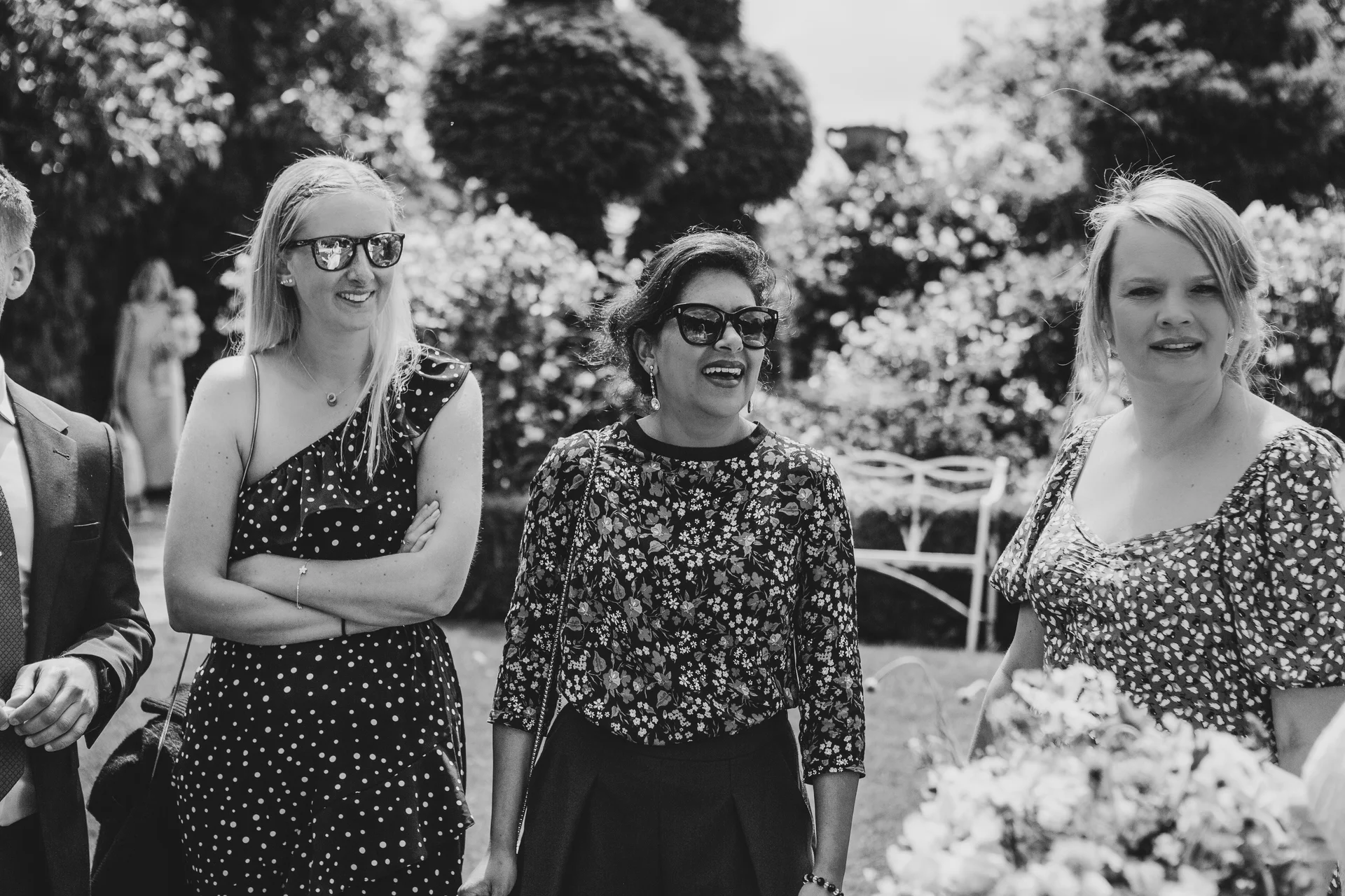 Three female wedding guests wearing sunglasses and patterned dresses laugh together outdoors in a sunlit English country garden.