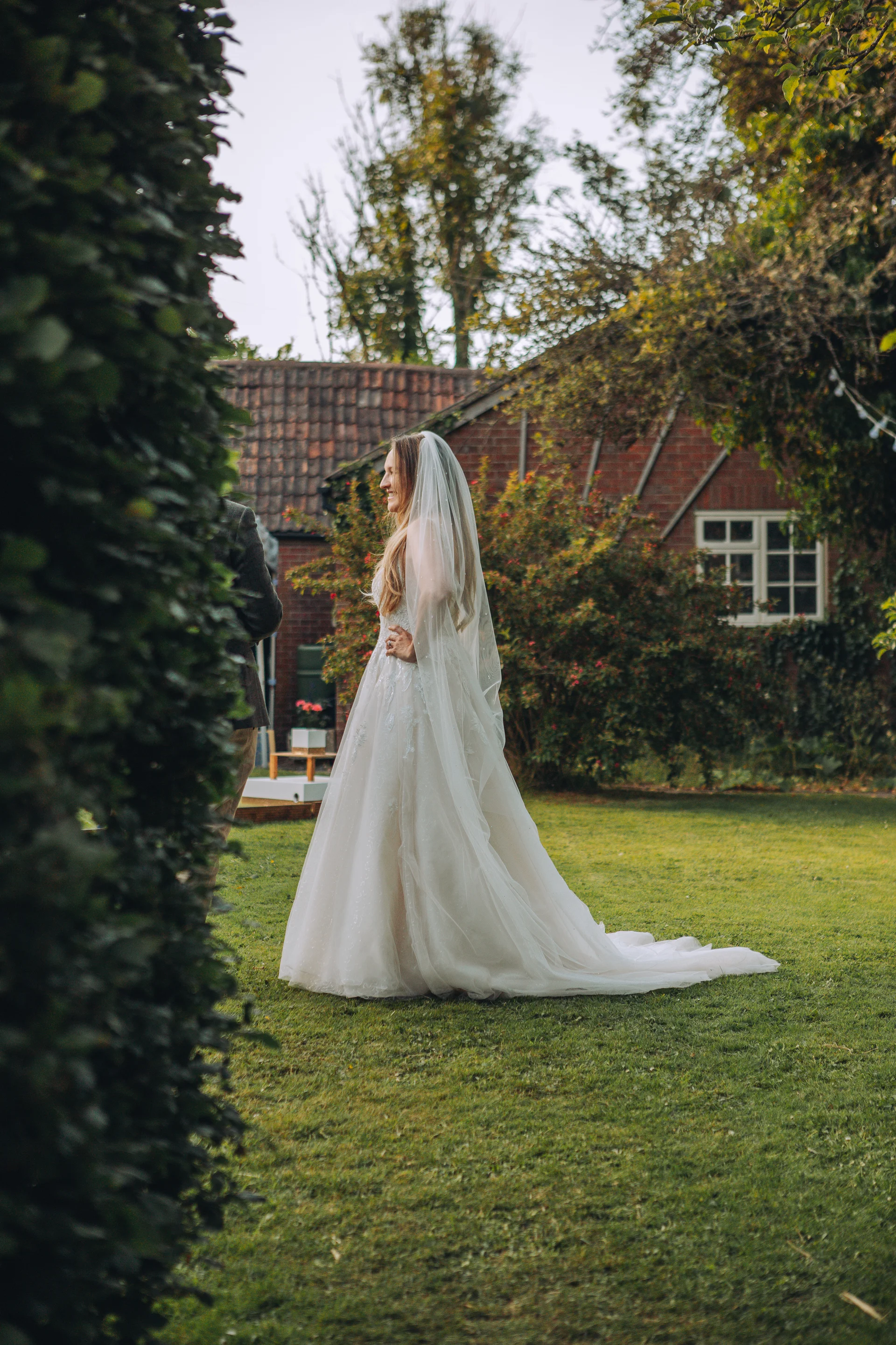 A smiling bride wearing an A-line wedding gown and chapel veil stands on a lush lawn in front of a rustic brick building during an outdoor portrait session.