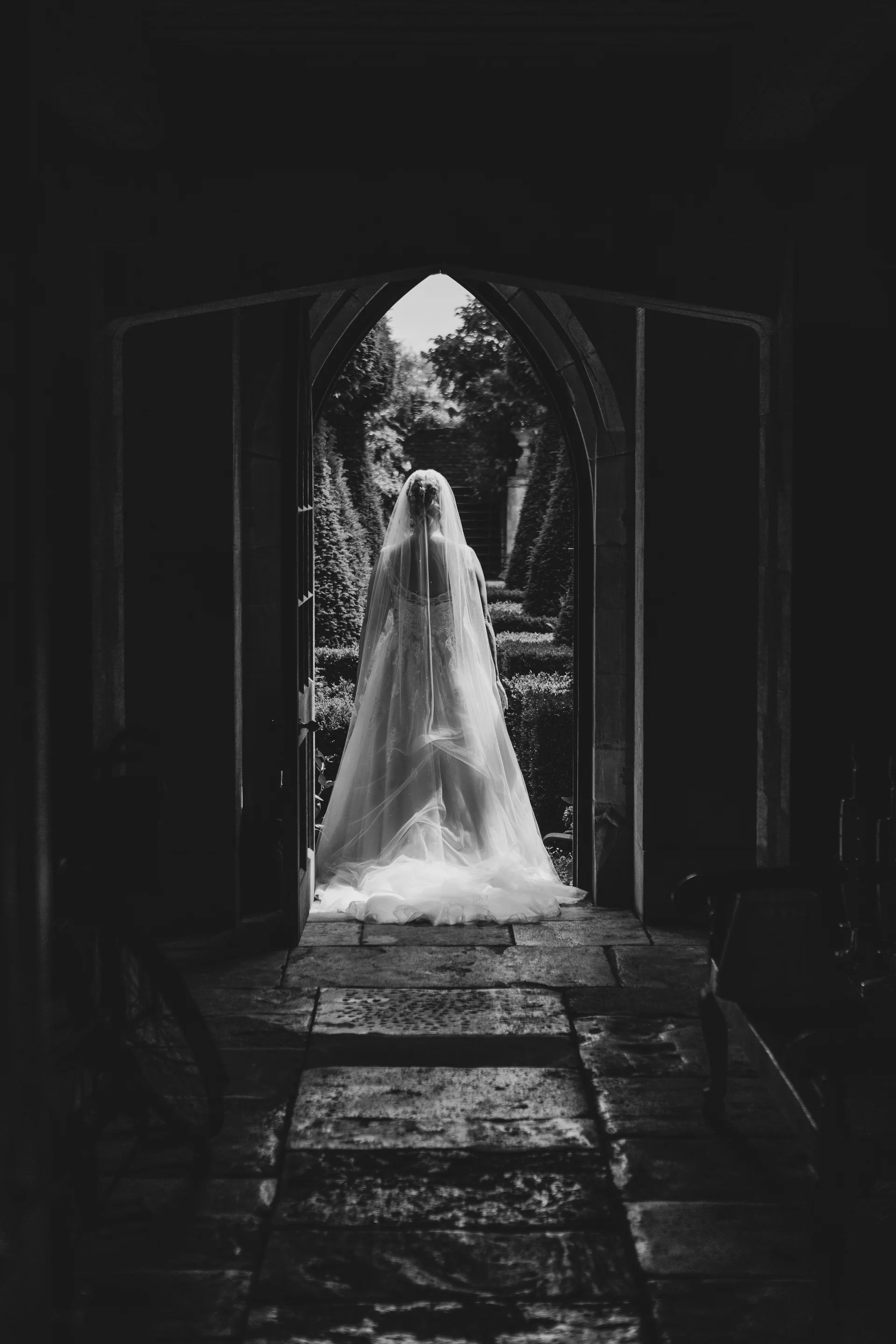 A bride viewed from behind stands framed in a historic stone doorway, wearing a classic a-line dress and long veil while looking out toward a lush courtyard.