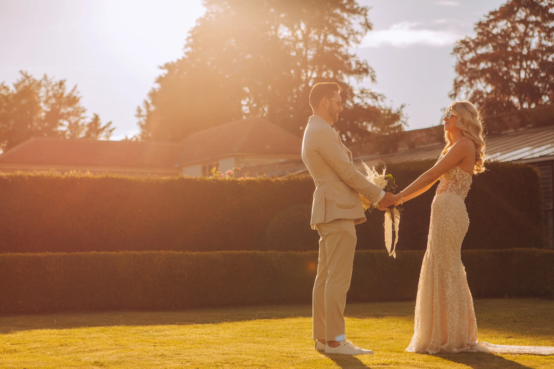 A stylish bride and groom holding hands and wearing sunglasses while standing on a sunlit lawn during golden hour.