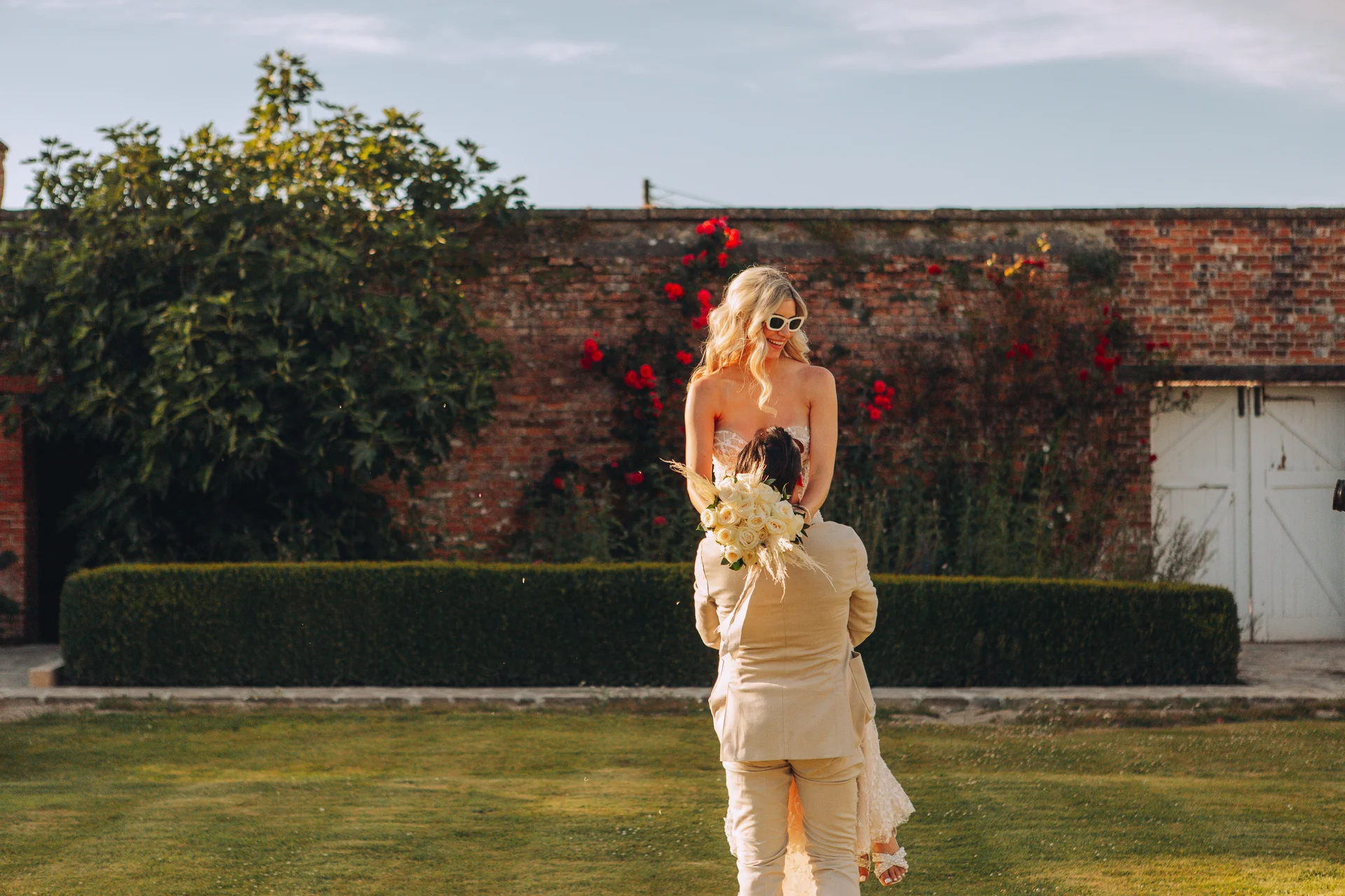 A groom joyfully lifts his bride in the air outdoors in a walled garden featuring a rustic brick wall, lush green lawn, and climbing red roses.