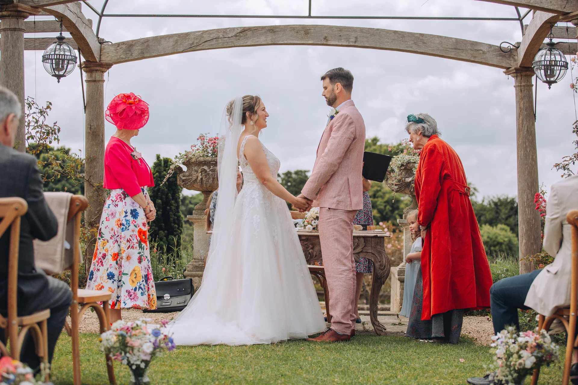 A bride and groom holding hands during an outdoor ceremony under a wooden pergola, surrounded by guests in colourful wedding attire.