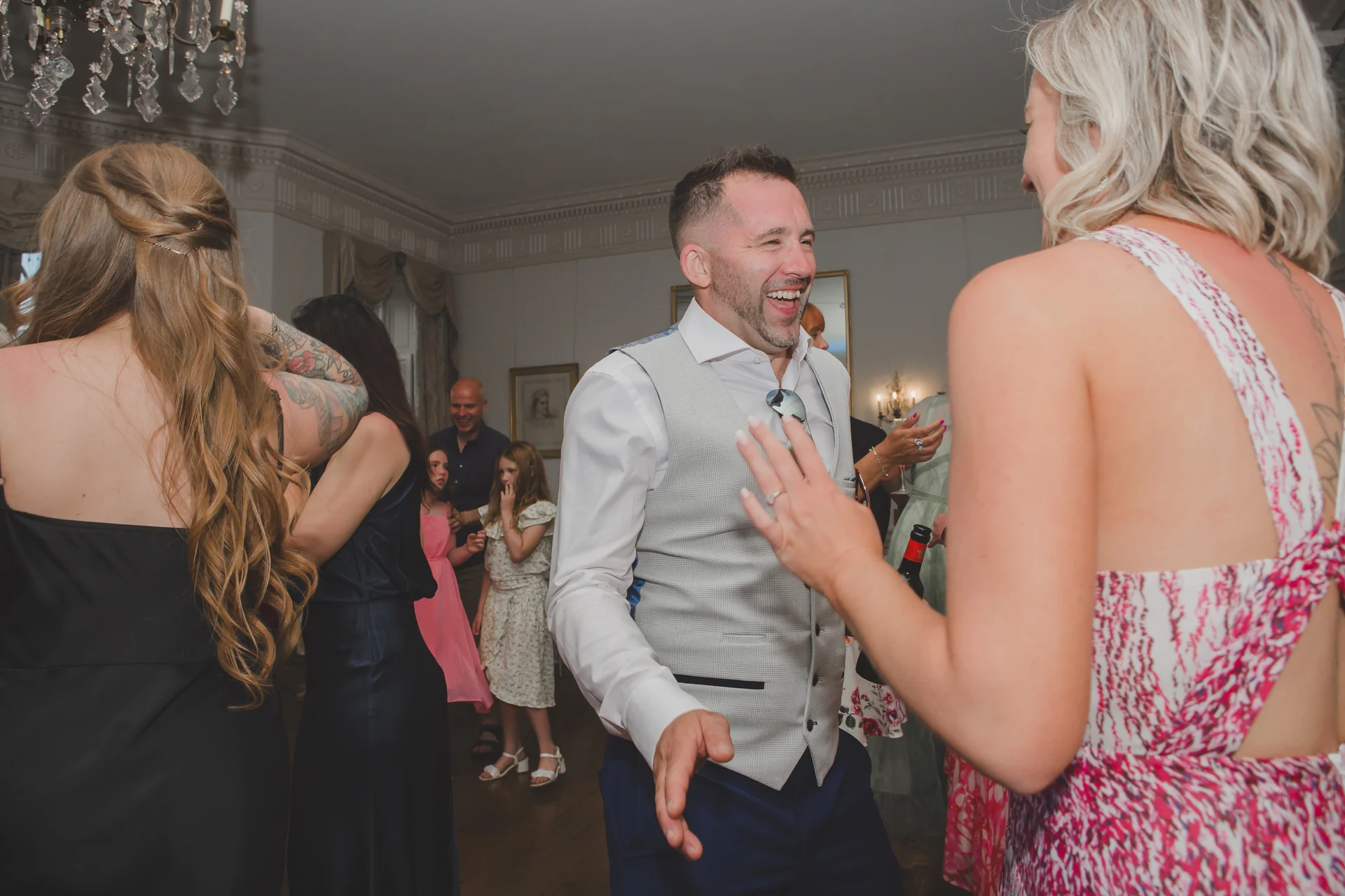 A groom laughs joyfully while dancing with guests on a lively evening reception dance floor in an elegant country house.