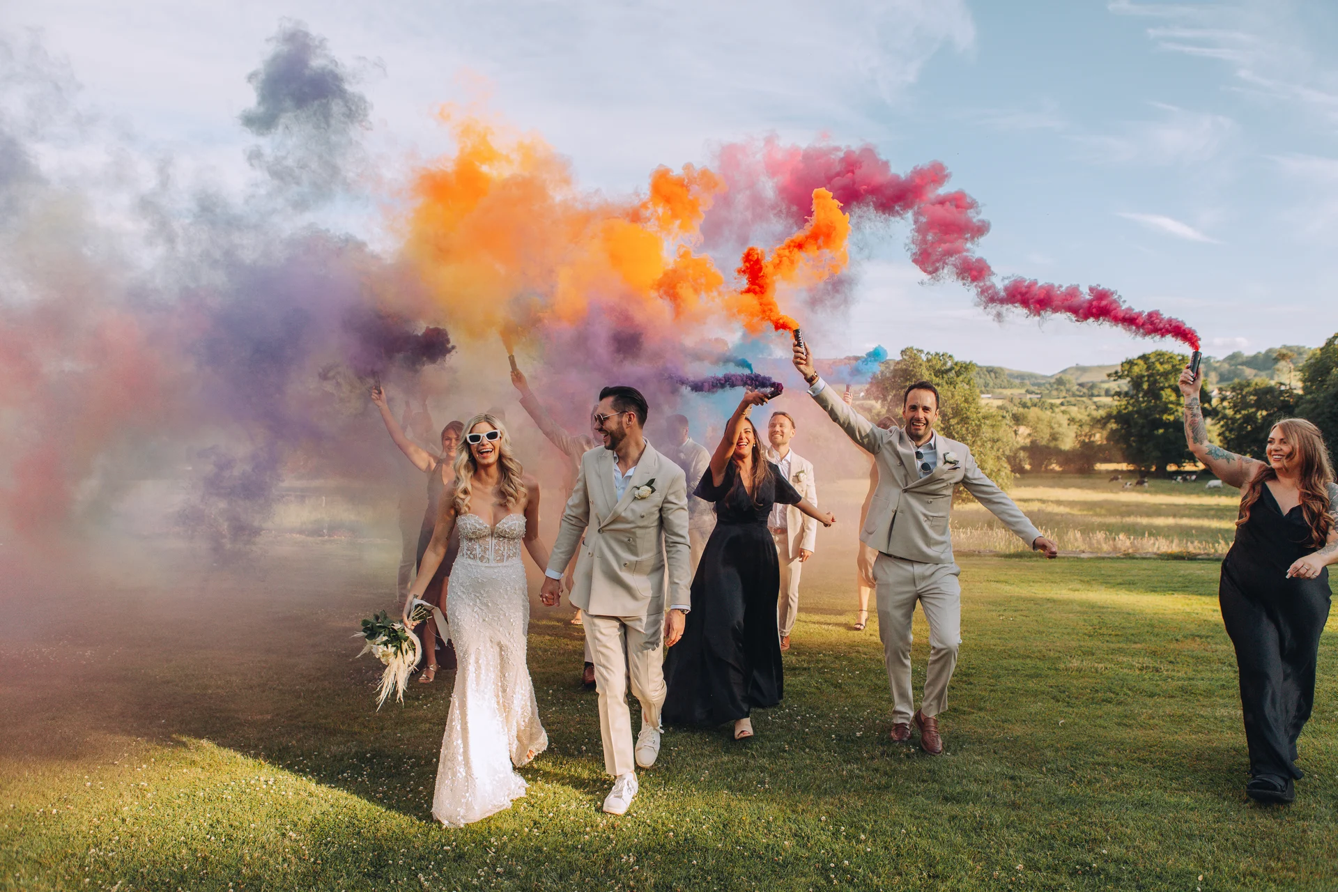 A bride and groom walk through a grassy field with their wedding party while holding colorful smoke bombs that create a vibrant cloud of smoke behind them.