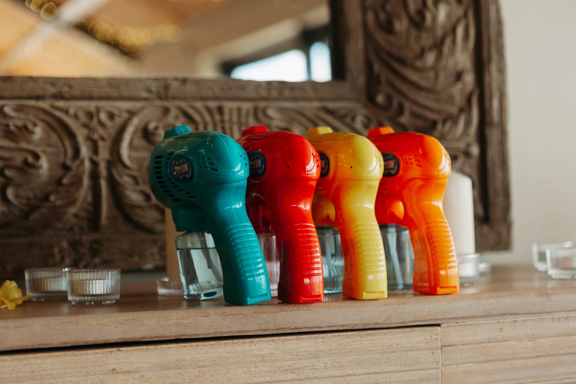 A line of bright, multi-coloured toy bubble guns sits on a wooden surface against a rustic carved mirror backdrop.