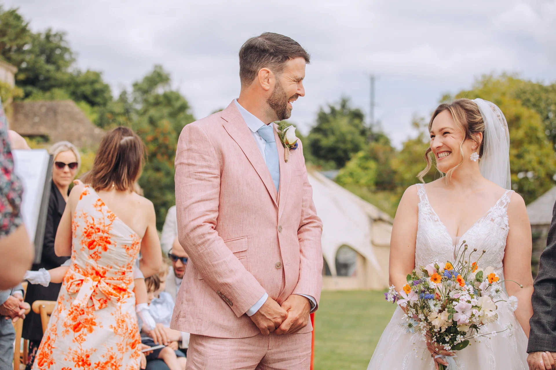A smiling bride and groom share a joyful moment during an outdoor wedding ceremony, with the groom wearing a pink suit and the bride holding a colourful wildflower bouquet.