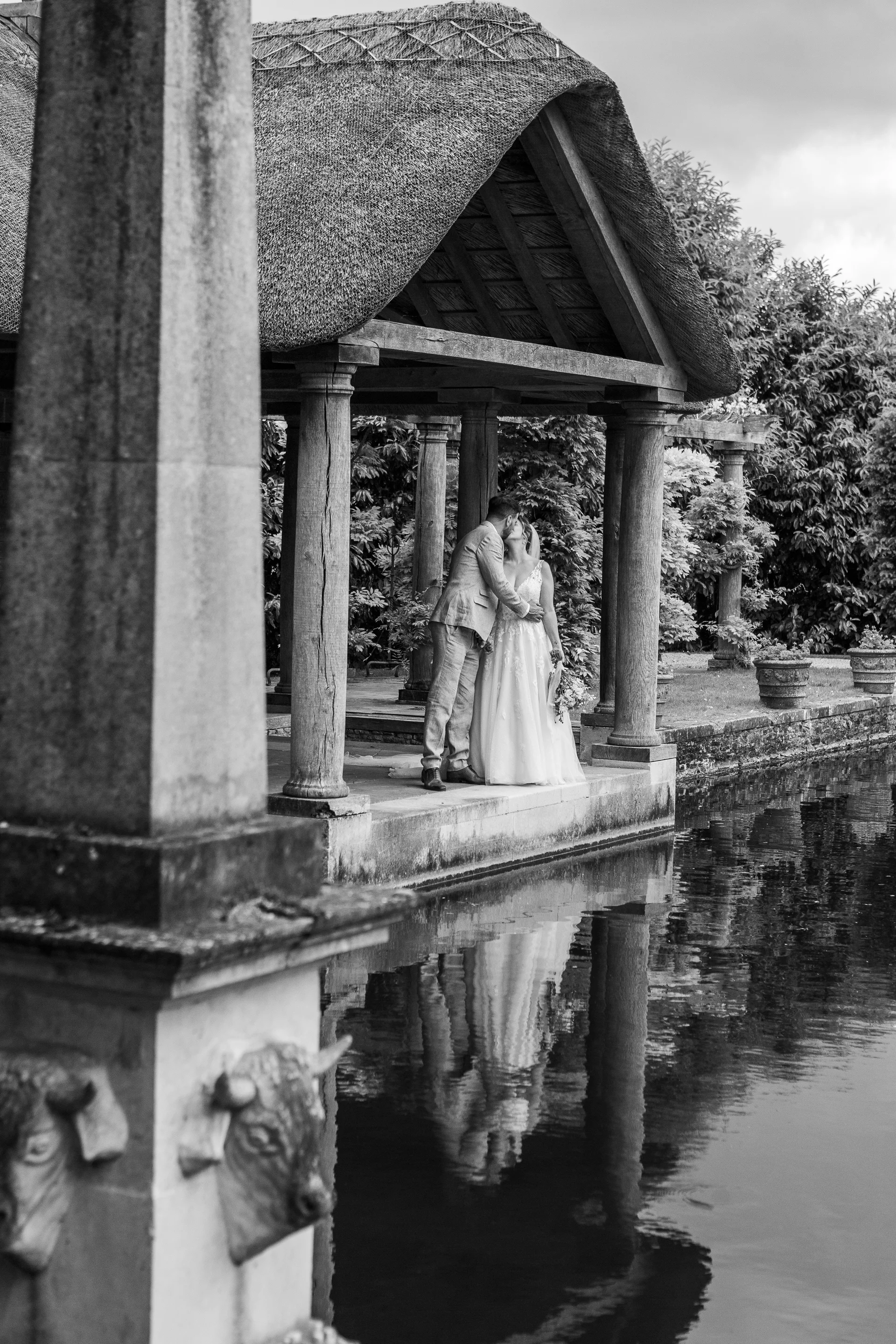A newlywed couple shares a tender kiss inside an open-air stone pavilion alongside a tranquil lake, creating a stunning reflection in the water below.