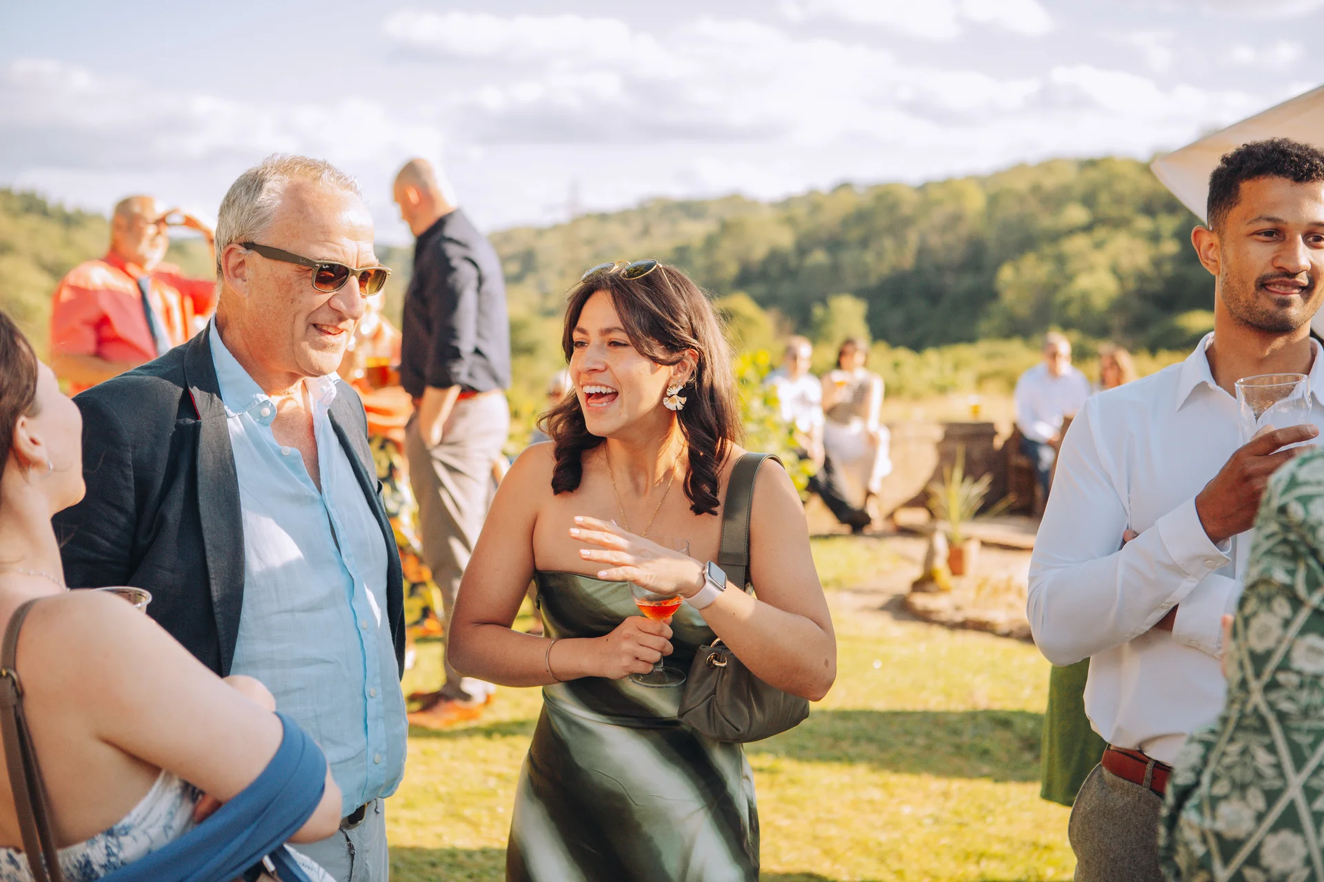 Guests laugh and chat together outdoors during a sunny wedding drinks reception.
