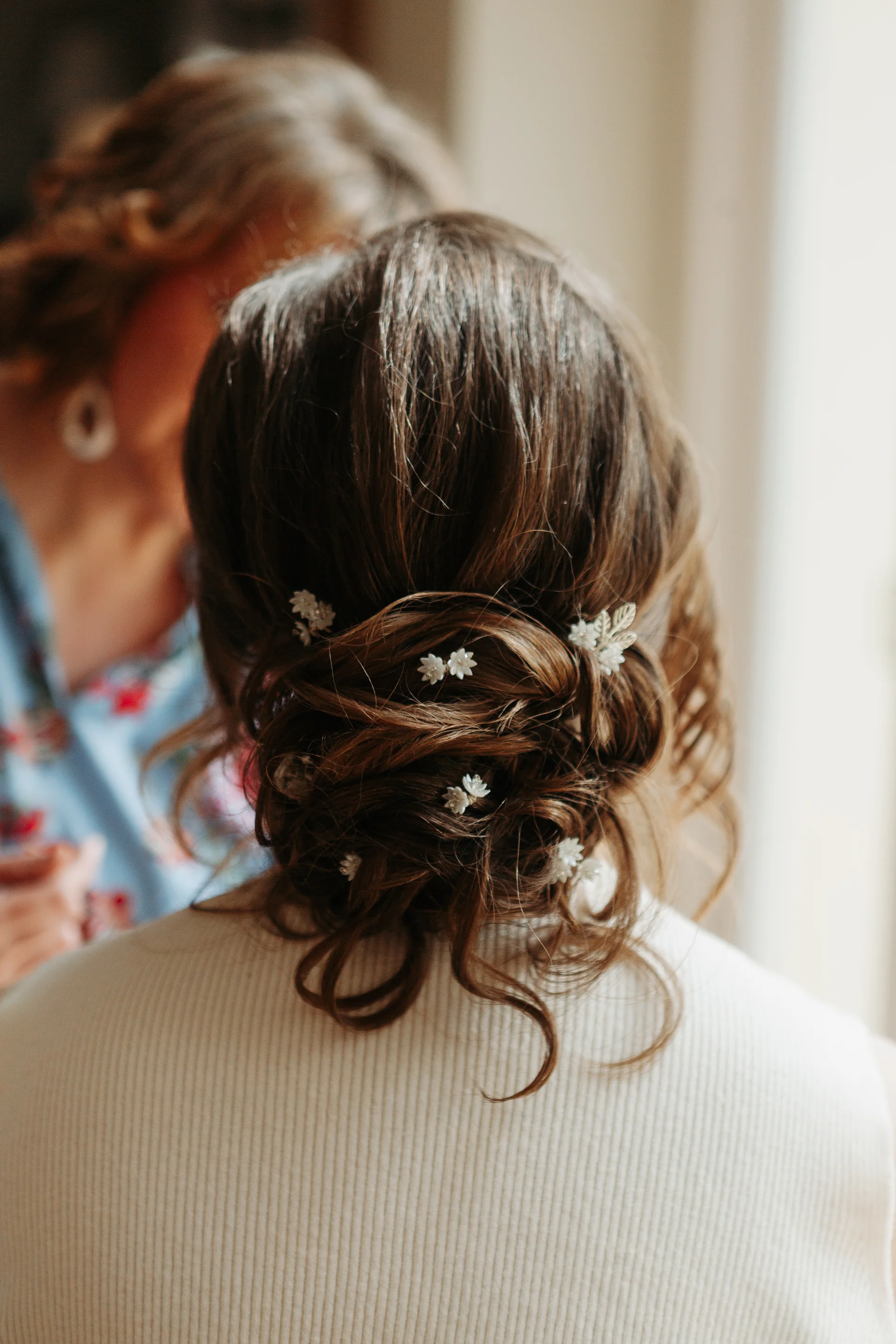 A close-up back view of a bride getting her hair styled into an intricate, textured low bun adorned with delicate floral hair pins during morning preparations.