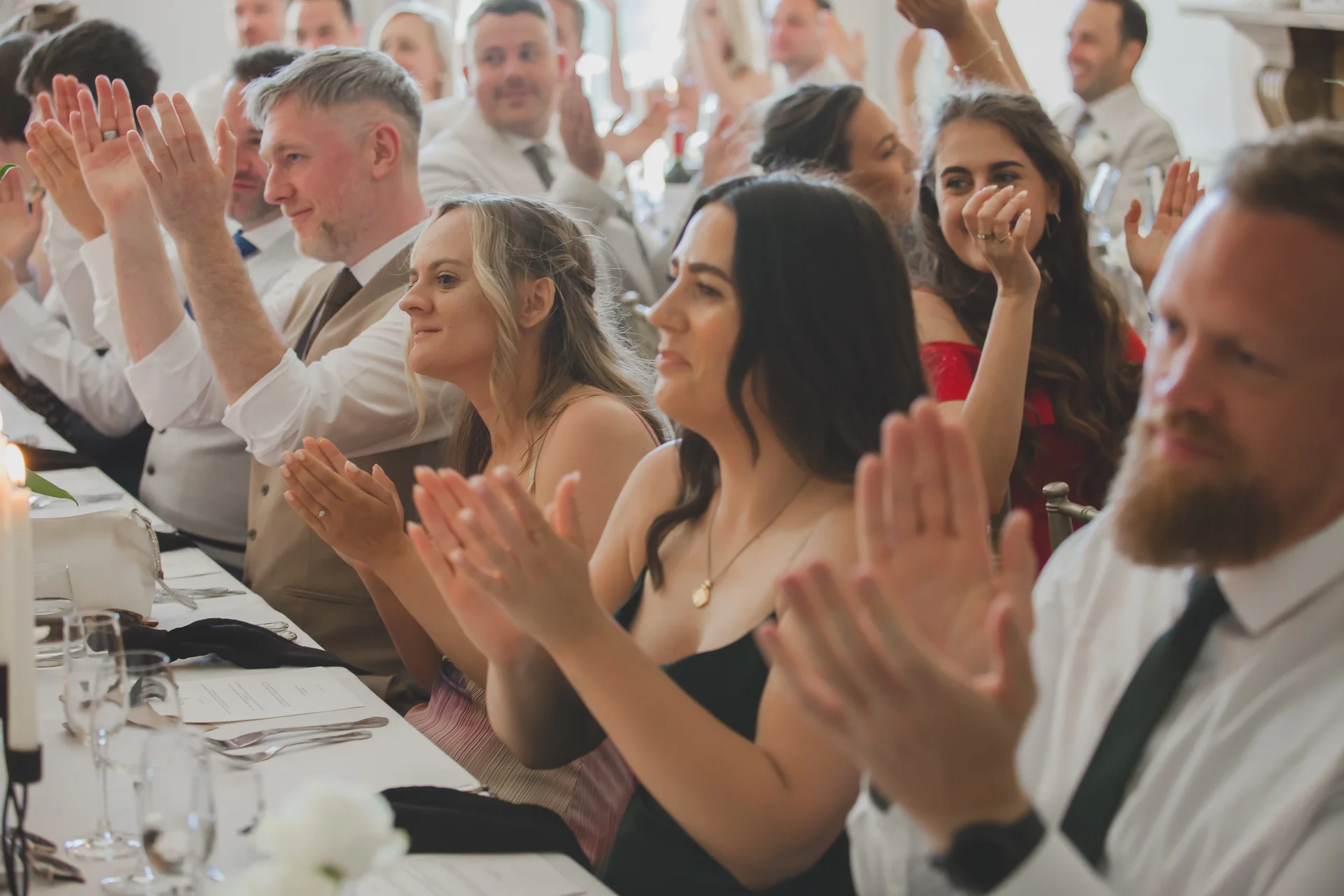 Wedding guests seated at a long banquet table clap and smile joyfully during speeches.