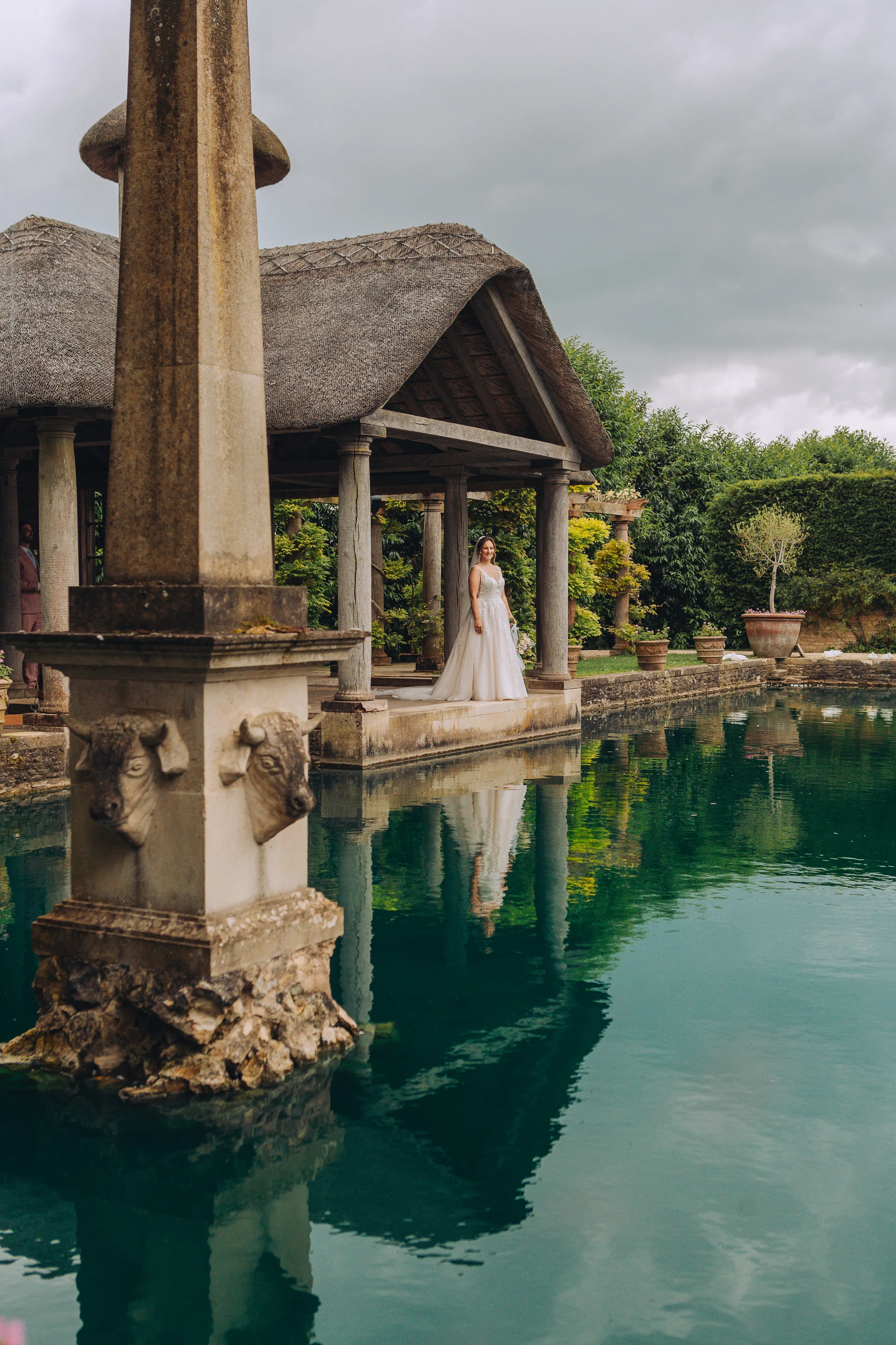 A bride stands gracefully near a tranquil ornamental lake under an overcast sky, with her reflection shimmering in the calm green water beside a stone structure.