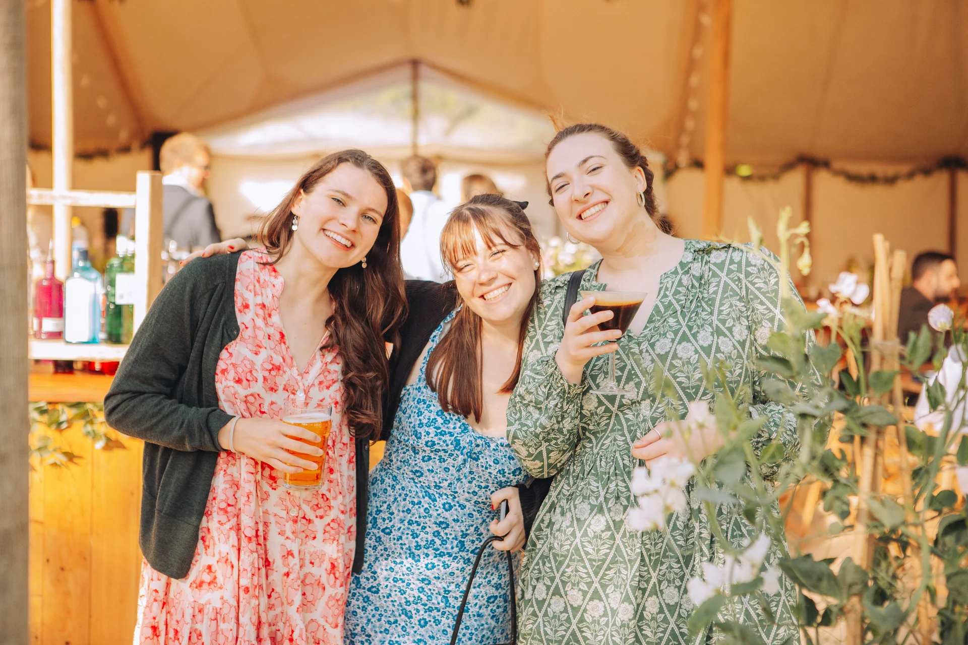 Three smiling wedding guests pose happily together inside a marquee during a drinks reception, holding drinks and wearing colourful patterned dresses.