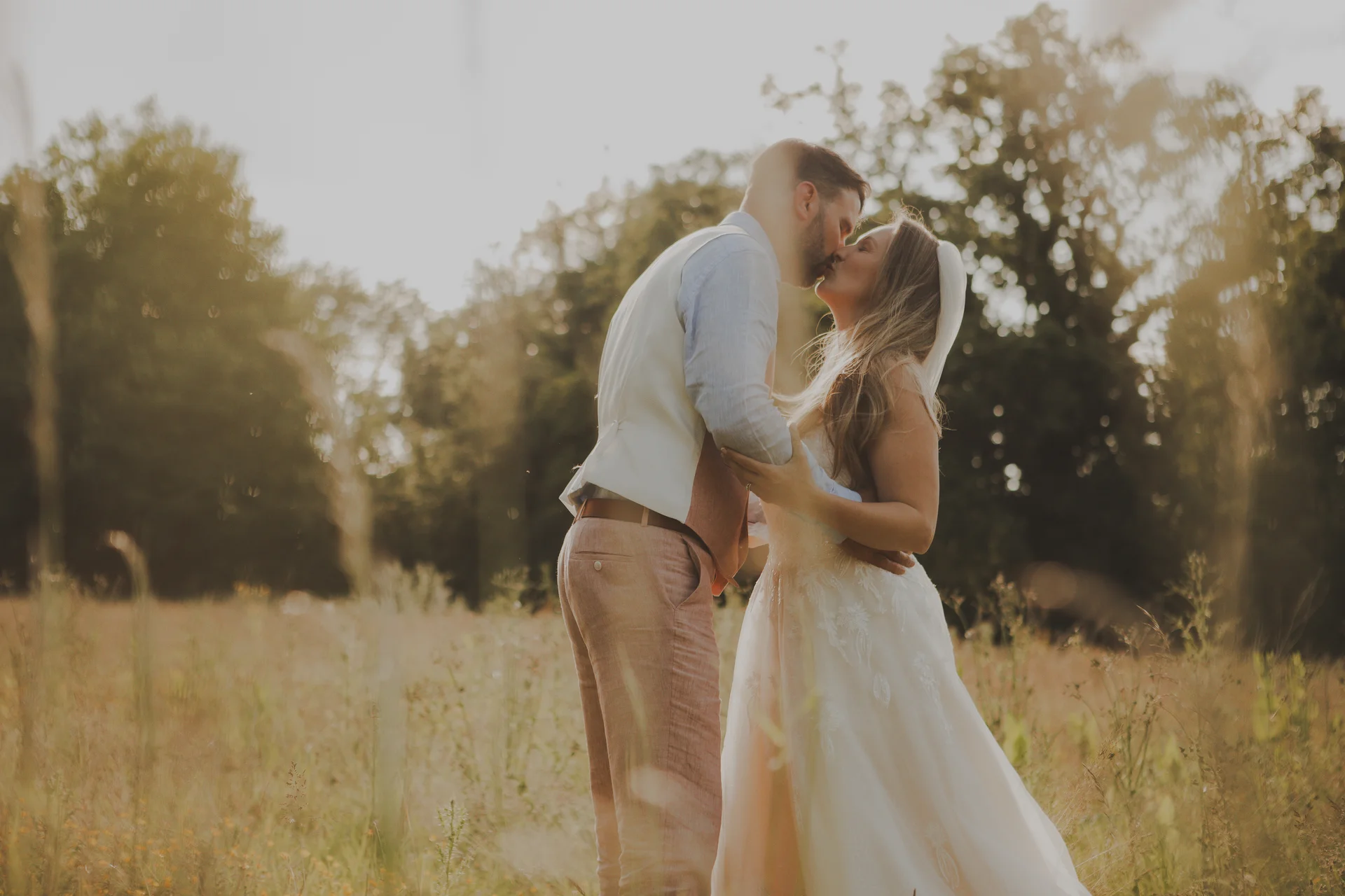 A newlywed couple shares a tender kiss in a sunlit meadow during golden hour, surrounded by tall grass and trees.