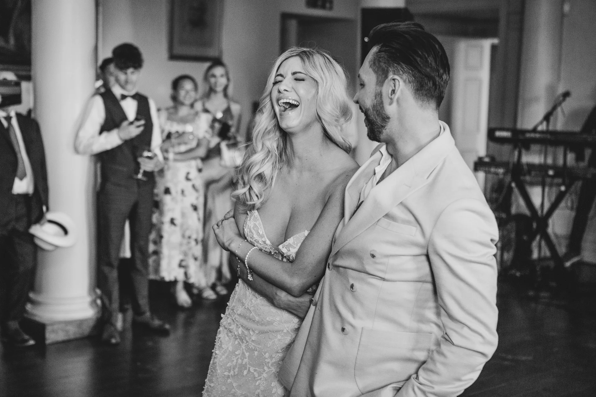 A newlywed bride and groom share a moment of pure laughter and joy together at their wedding reception, surrounded by smiling guests.