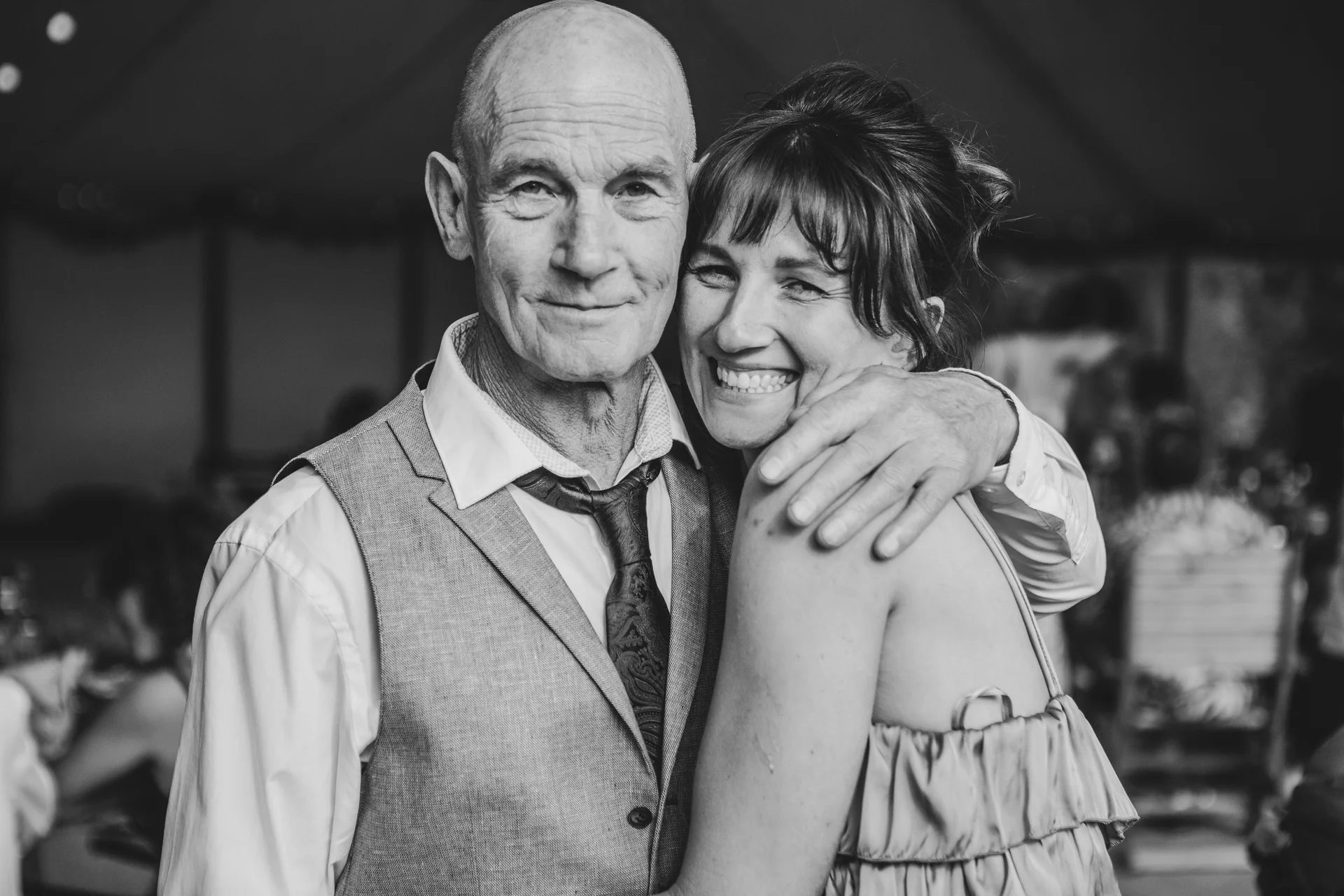 An older couple in formal attire share a joyful embrace and laugh together during a wedding celebration.