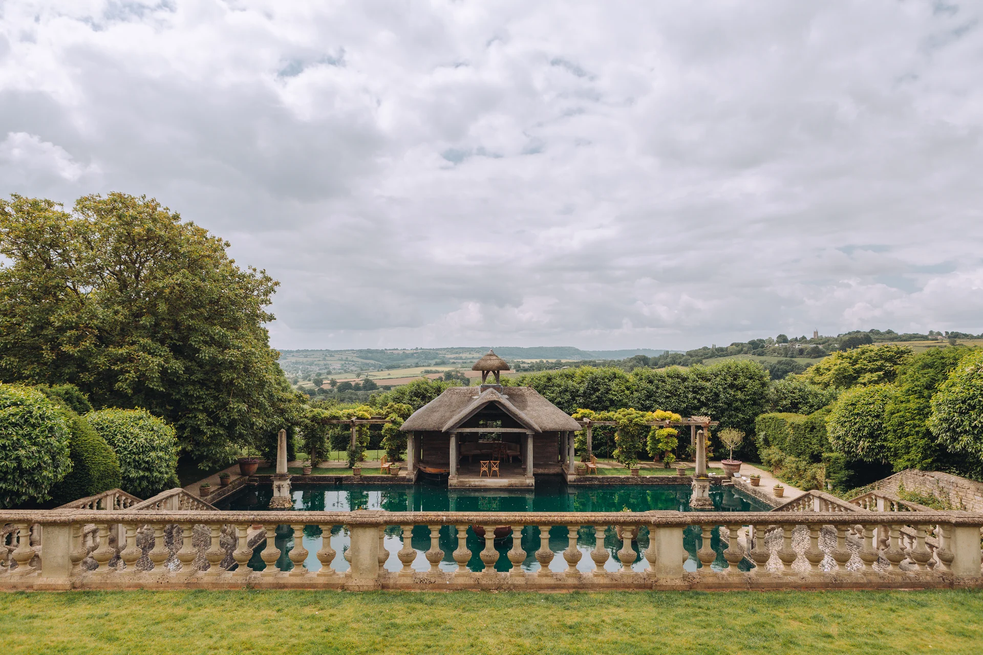 A sweeping outdoor view of a country house wedding venue featuring a thatched pavilion by a tranquil lake under an overcast sky.