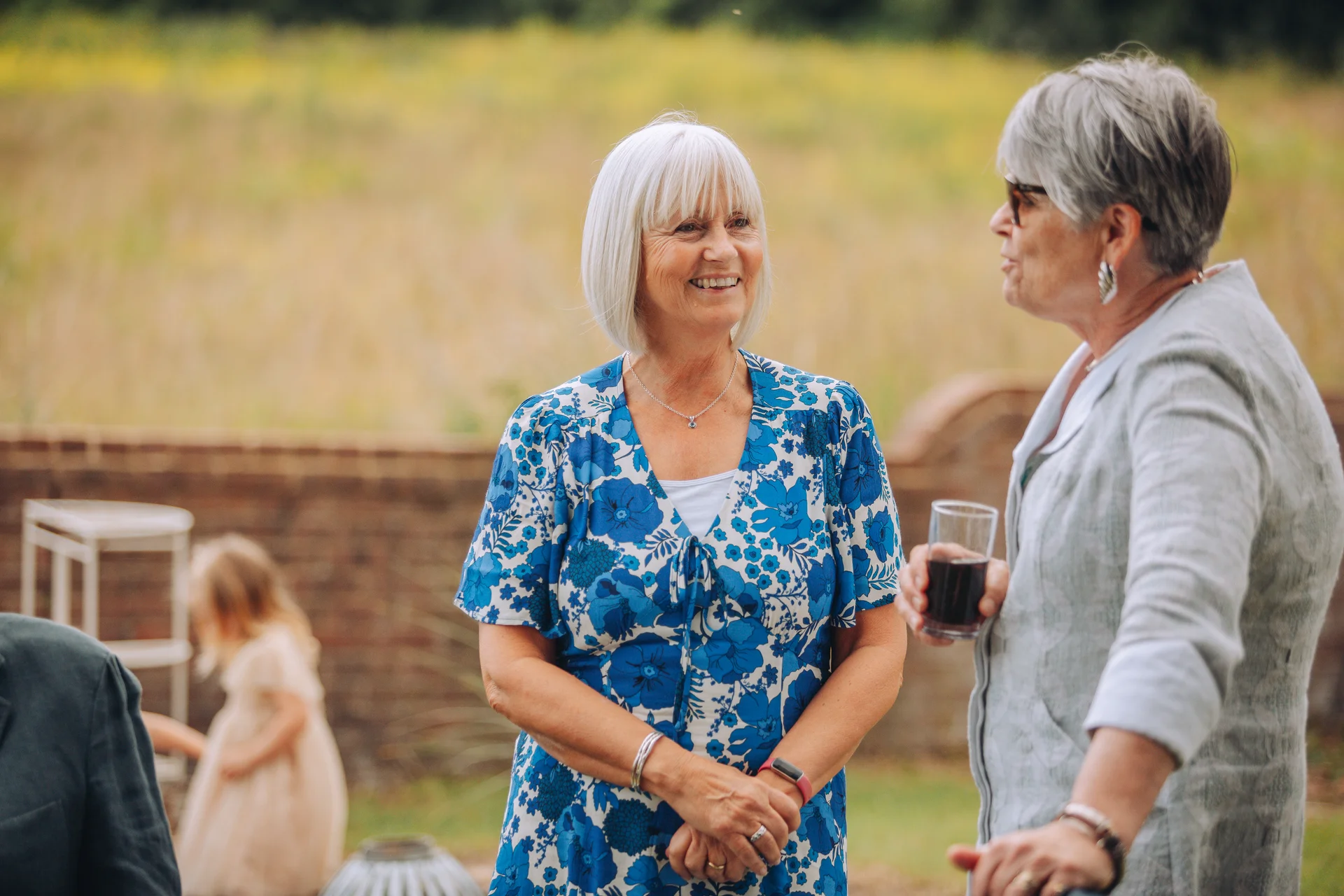 Two older female guests smiling and chatting together outdoors at a wedding drinks reception.