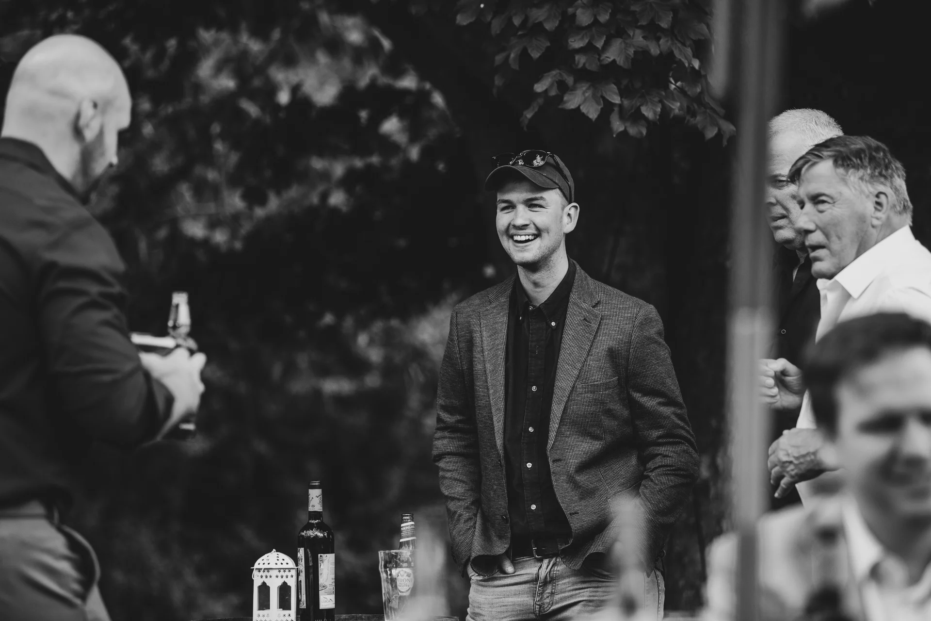 A candid black and white photograph captures wedding guests laughing and mingling outdoors during the drinks reception near a table with bottles and a lantern.