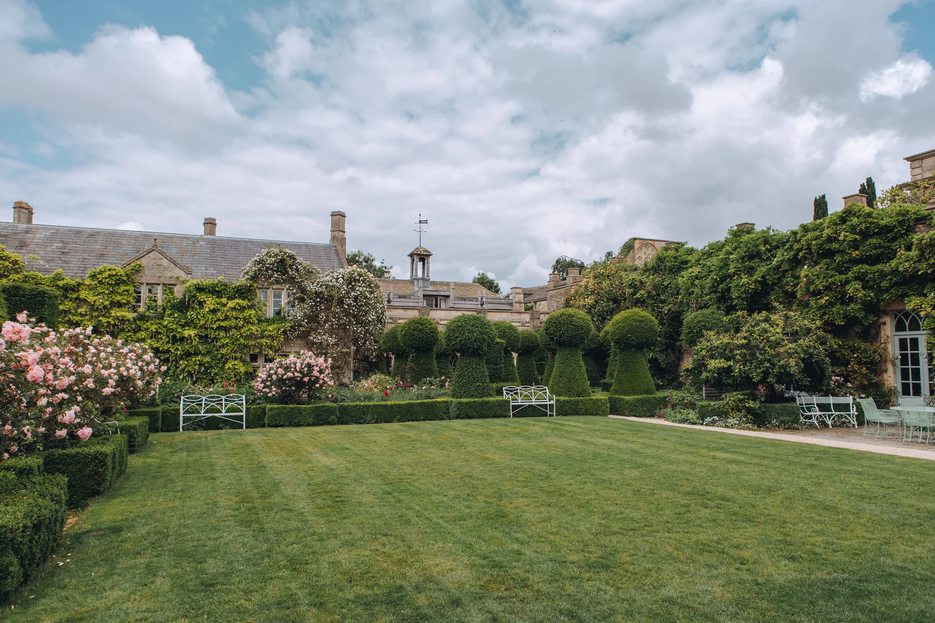 An expansive manicured lawn of a country manor house wedding venue features trimmed topiary trees, climbing roses along stone walls, and white decorative garden benches under an overcast sky.