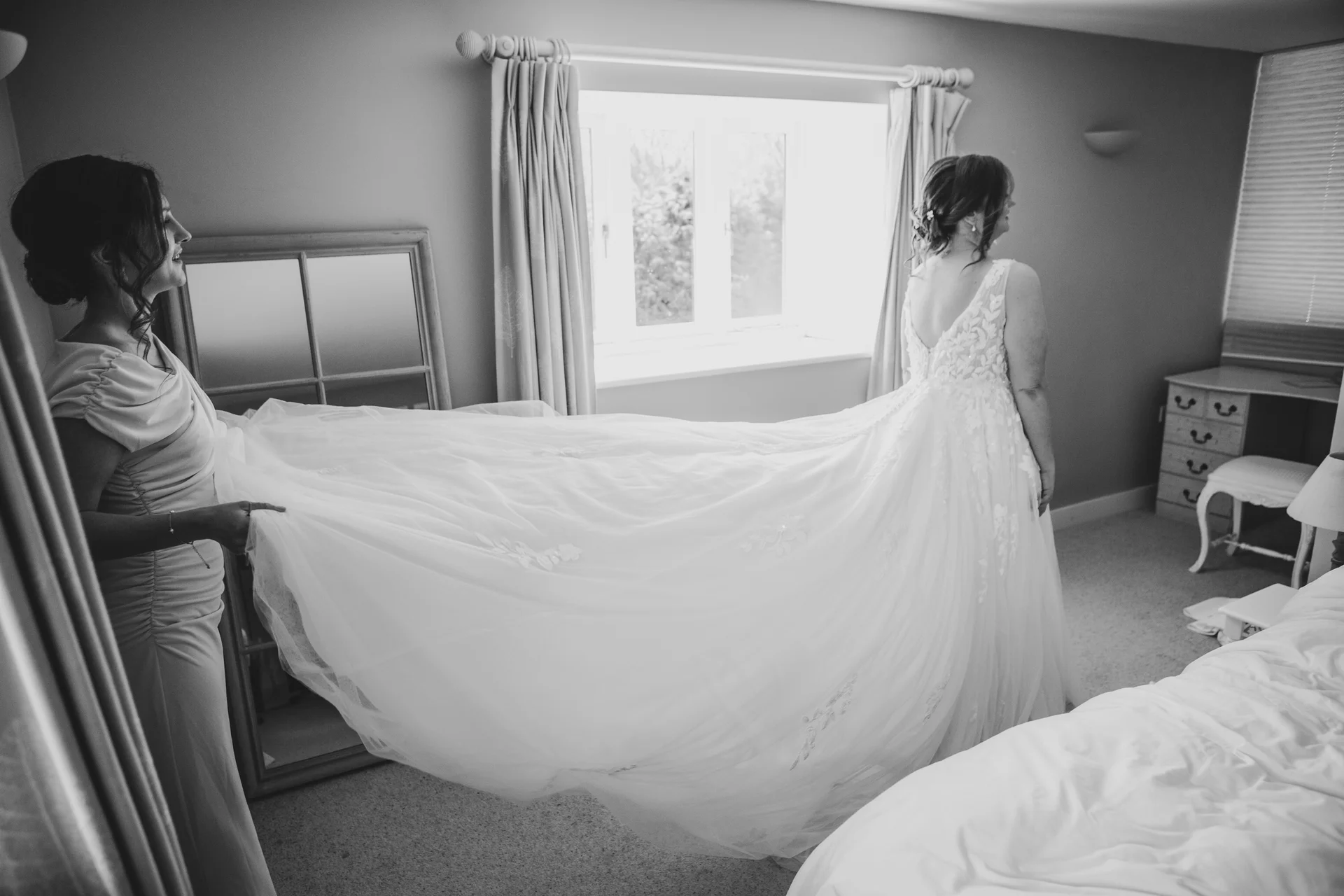 A black and white photograph captures a bridesmaid helping the bride adjust her long, flowing wedding dress train inside a bright bedroom.