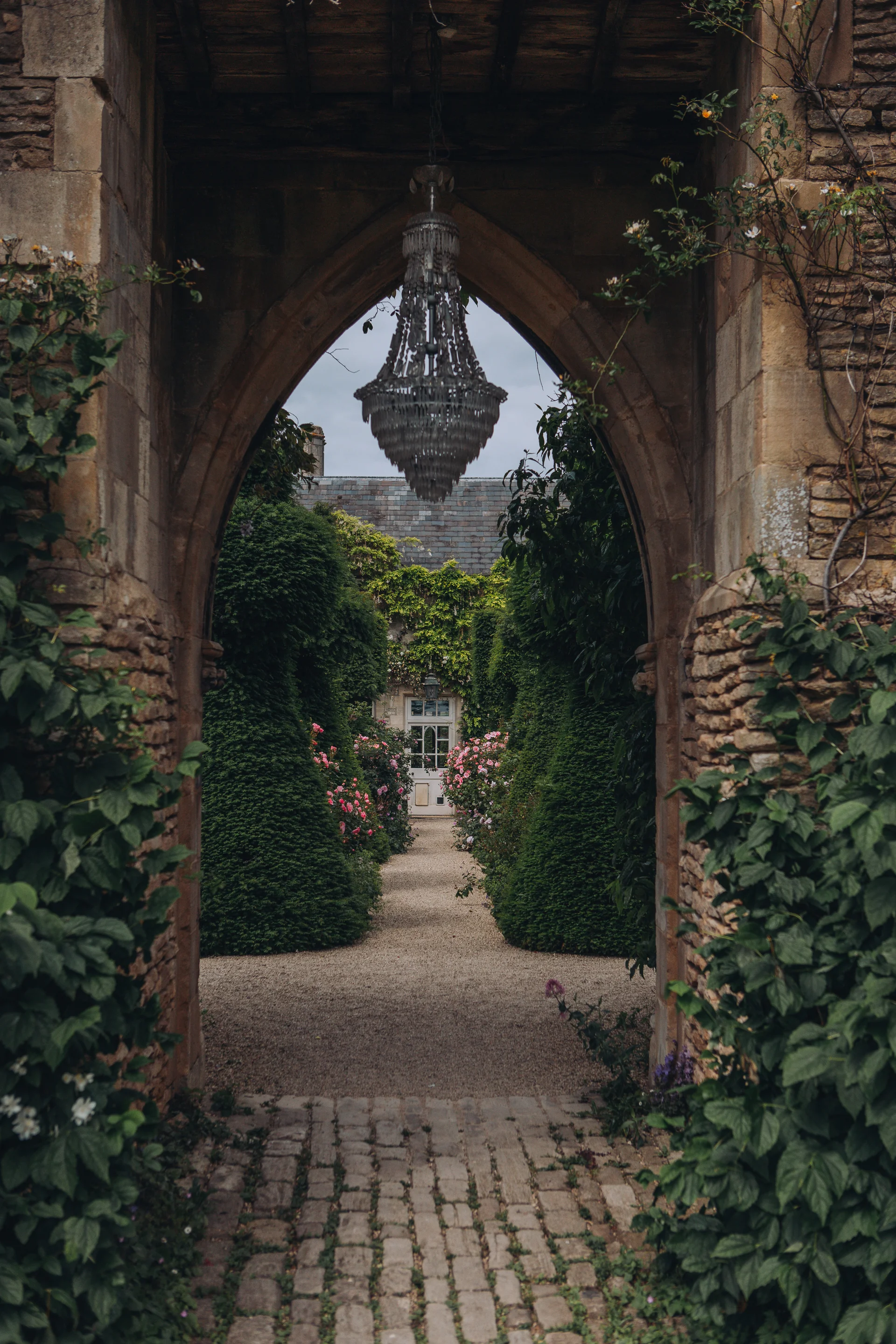 A rustic stone archway frames a scenic, manicured pathway lined with topiary bushes, blooming roses, and a hanging crystal chandelier.