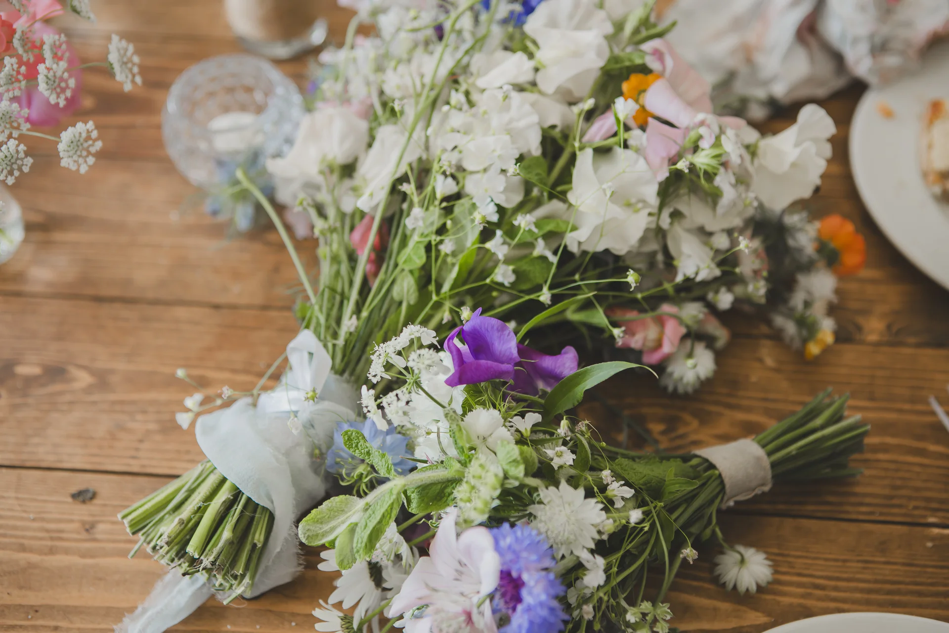 A close-up flat lay shot of a wild, meadow-style wedding bouquet featuring sweet peas and cornflowers tied with a delicate silk ribbon resting on a rustic wooden table.